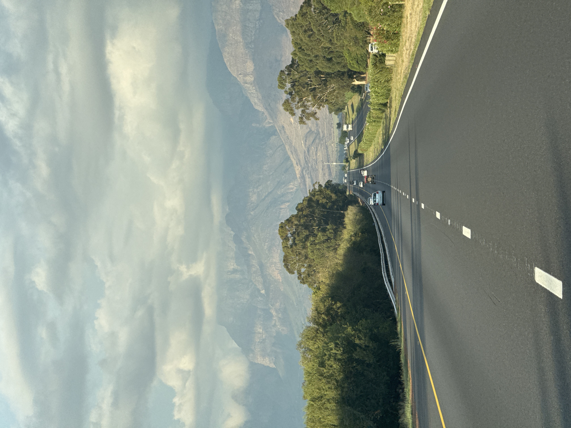 The image depicts a road in Drakenstein Nu, South Africa. A paved road with painted lane markings stretches into the distance. Several vehicles are traveling along the road. The road is bordered by green vegetation and trees. In the background, there is a mountain range obscured by haze or clouds. The sky is overcast.