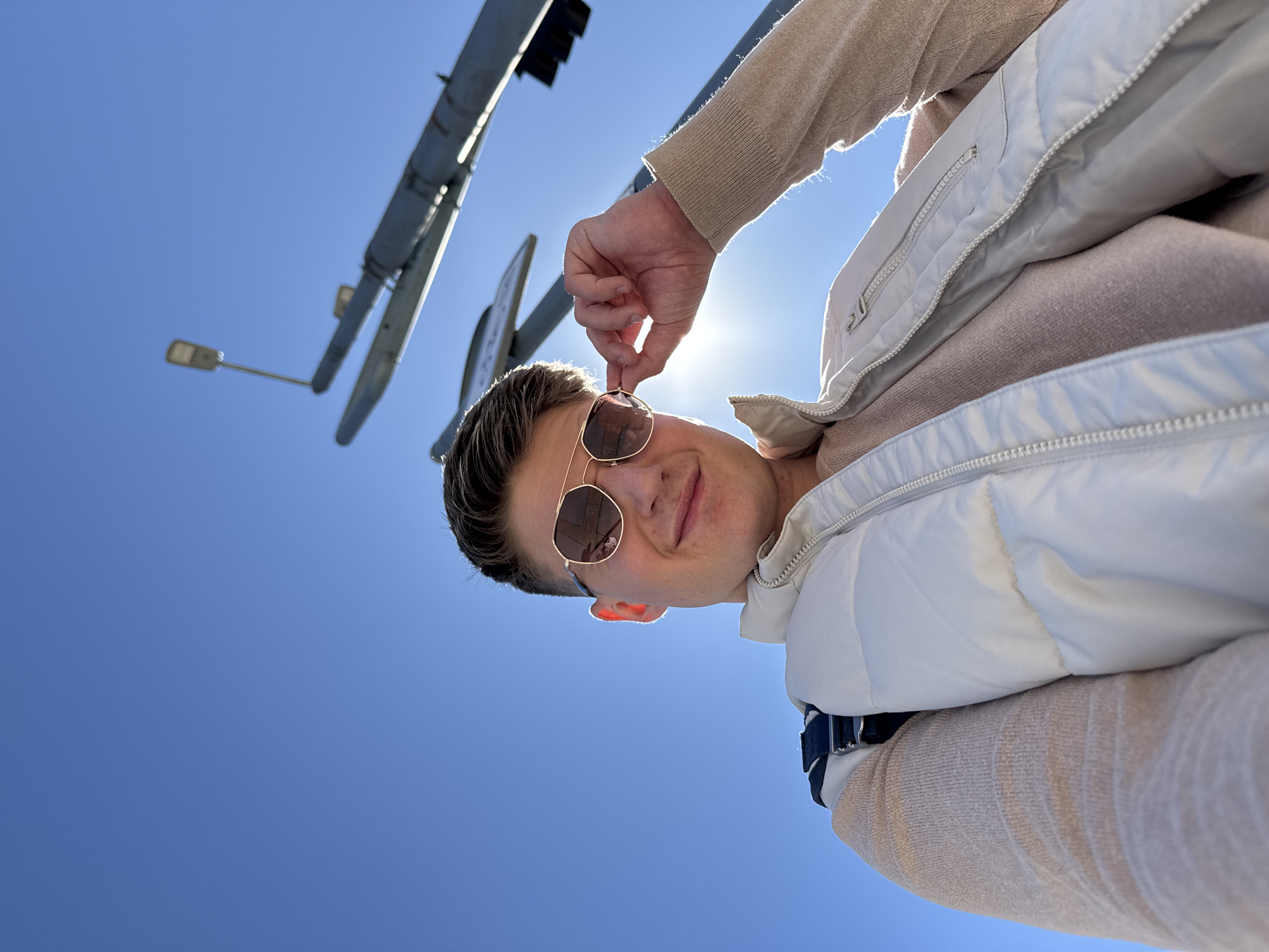 The image is taken from a low angle, looking up at a man wearing sunglasses, a light beige sweater, and a white vest. He is holding the frame of his sunglasses with his right hand. Above and behind him, there is a traffic light and a street lamp against a clear blue sky. The exact location within Plovdiv, Bulgaria, is not discernible from the image.
