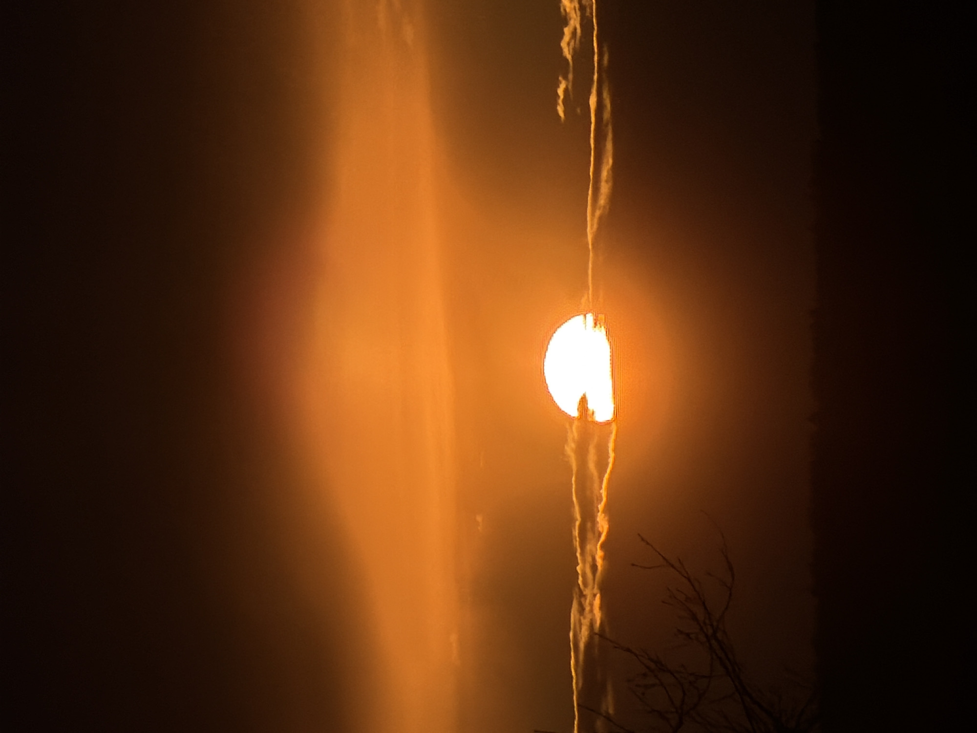 The image, captured in Chaikovsky, Russia, presents a striking sky scene dominated by warm orange and amber tones, suggesting either sunrise or sunset. A brilliant, D-shaped celestial body, likely the sun, is prominently featured on the right side of the frame. It emits intense white light from its center, surrounded by a soft, diffused orange glow that illuminates the hazy atmosphere. A distinct, thin, and elongated wispy cloud or contrail extends vertically, piercing through the bright D-shaped sun and continuing upwards across the frame. To the left of this central vertical feature, broader, softer clouds are visible, also bathed in the warm light. Towards the bottom right, subtle silhouettes of bare tree branches can be seen against the glowing sky. The far right and left portions of the image are considerably darker, providing a stark contrast to the bright, luminous center. No people or visible text are present in the image. The scene evokes a sense of natural beauty and dramatic light.