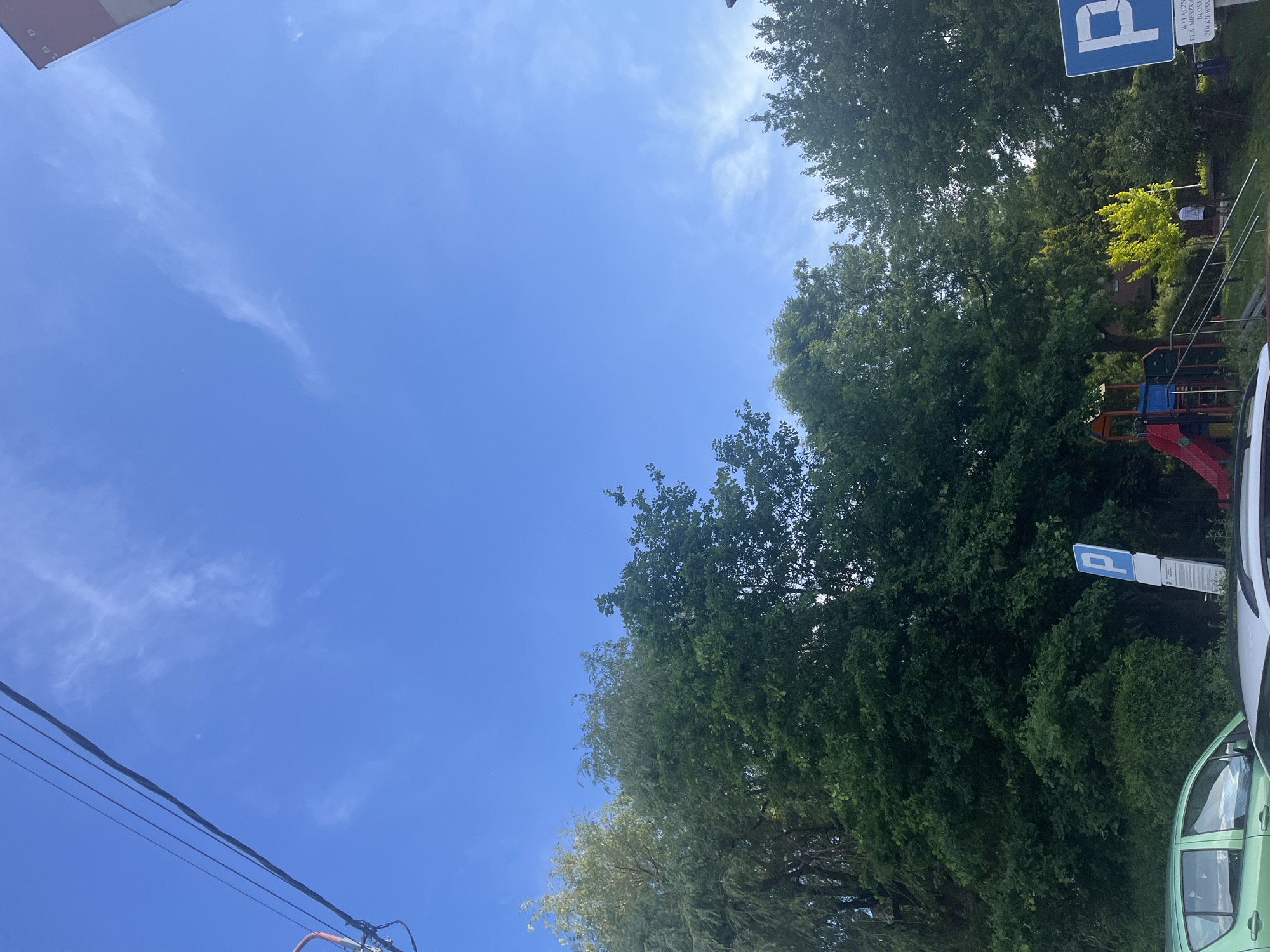 The image presents a view of a park or recreational area in Unknown. Dominating the frame is a clear blue sky with wispy clouds. Below, lush green trees form a natural barrier. A blue parking sign is visible among the trees. A playground with a red slide is present. Two vehicles are parked nearby. Power lines are also visible.