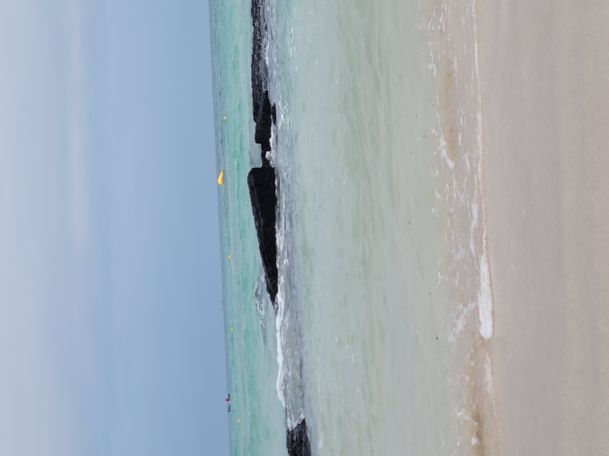 The image shows a serene beach scene with calm, shallow water lapping onto a sandy shore. The water transitions from a pale, almost white hue near the shore to a clearer turquoise further out. In the middle ground, dark, jagged rocks break the surface of the water, with gentle waves creating white foam as they wash around them. In the distance, there are a few yellow buoys floating on the water, and a small boat with a red sail can be seen on the far left. The sky is overcast with a soft, muted blue. The overall impression is one of tranquility and natural beauty, typical of a coastal area. While the image itself doesn't explicitly identify the location as Jeju, South Korea, the description of such a beach with volcanic rocks is consistent with many coastal areas on the island.