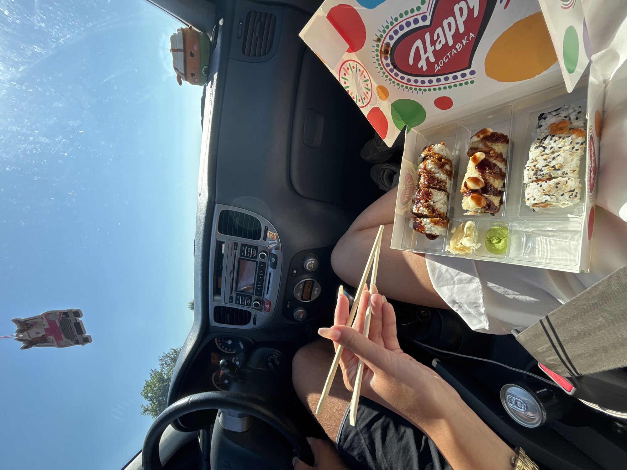 The image shows the interior of a car, with a focus on a meal of sushi. A person's hands are visible, holding chopsticks over a take-out container of sushi. The container is plastic and divided into sections, with various types of sushi rolls, pickled ginger, and wasabi. A can of soda is also visible in the car. The car appears to be parked or in slow motion on a sunny day, with the sky visible through the windshield. The dashboard and steering wheel of the car are visible. There are also two car air fresheners hanging from the windshield. The overall scene suggests a casual meal being enjoyed in a vehicle. The location is identified as the city of Chirpan, Bulgaria.