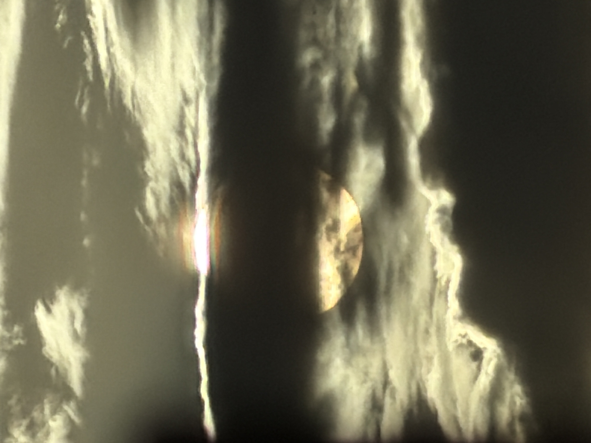 The image depicts a partially obscured sun seen through a cloudy sky. The sun, a glowing orb, is visible in the lower center of the frame, with its upper portion hidden behind dark clouds. Lighter, wispy clouds surround the sun and extend towards the top of the image, catching and diffusing the sunlight in a yellowish-white hue. The overall lighting suggests either sunrise or sunset. The scene is a natural landscape and does not contain any identifiable urban elements or people. The location of Krasnyi Krym, Russia, is not visually evident from the provided image.