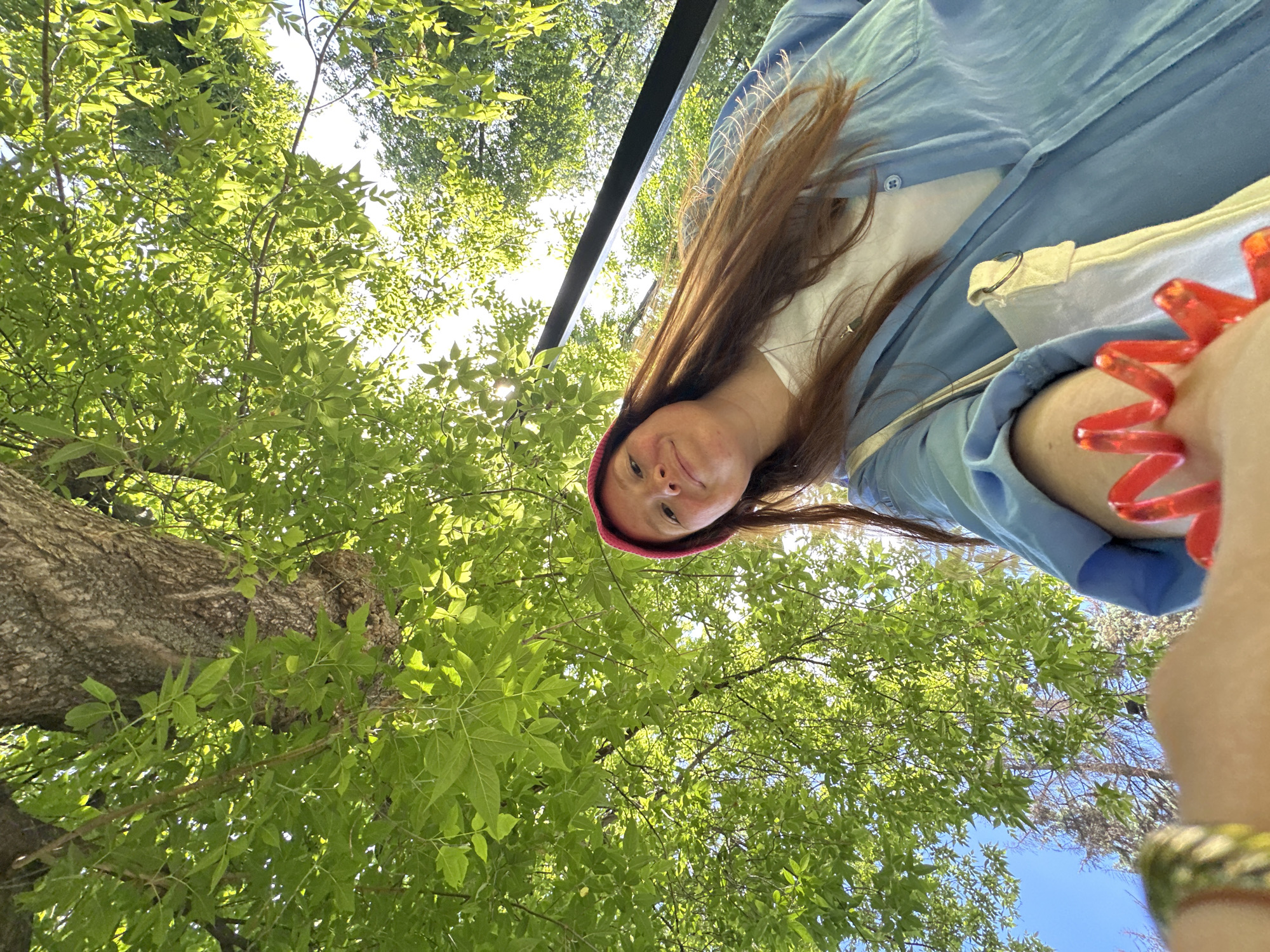 The image, rotated ninety degrees clockwise, presents a low-angle perspective looking upwards through a tree canopy towards a person. The main subject is a person, likely a woman, with long, straight brown hair, wearing a red beanie. She is looking directly at the camera with a calm, slightly smiling expression. She wears a light blue button-up shirt over a white top. Visible on her right arm (left side of the frame) is a translucent red, spiky bracelet, and a patterned yellow/gold bracelet on her left wrist (bottom right).
The background is filled with vibrant green leaves from a tree canopy, with bright sunlight filtering through, creating a dappled effect. A thick tree trunk is visible on the left side of the frame, extending upwards. Patches of bright white light and clear blue sky are seen through the foliage. A dark, straight, horizontal bar, possibly part of an overhead structure like a bridge or pergola, crosses the upper middle portion of the image, behind the person's head.
The setting is outdoors, likely a park or natural area, on a bright and sunny day. The low camera angle suggests the person is being photographed from below, possibly taking a selfie from a supine position or holding the camera high. The overall mood is relaxed and natural. There is no visible text. The location context provided is Nizhny Novgorod, Russia.