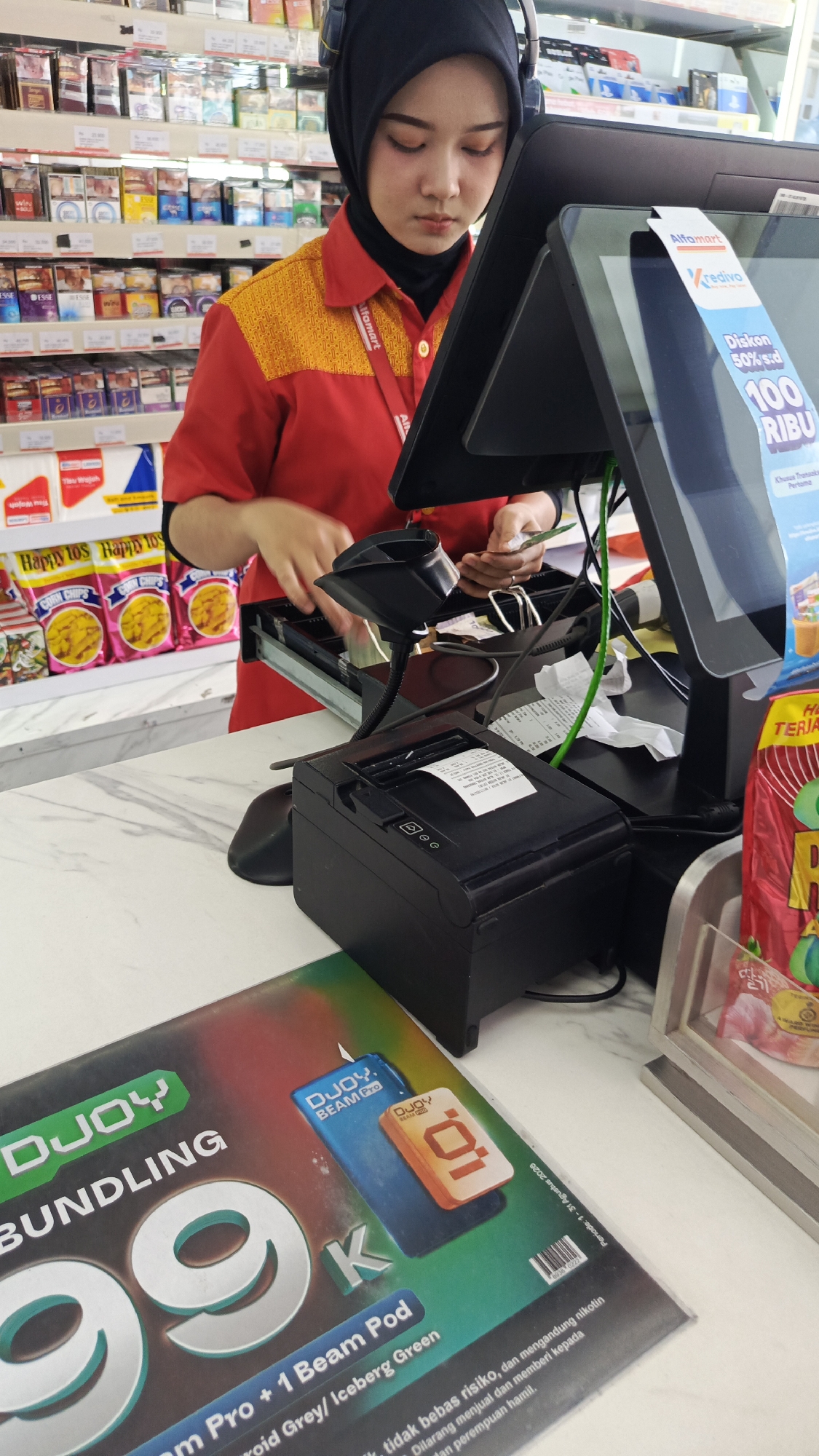 A young woman wearing a red uniform with yellow accents and a black hijab is operating a point-of-sale system at a checkout counter. She is focused on a computer screen and handling cash. A receipt printer is visible in front of her, along with various products on shelves in the background, suggesting a retail environment. The promotional material on the counter indicates a sale for a "DJOy Bundling 99K" which includes a "Beam Pro + 1 Beam Pod". The overall setting appears to be a convenience store or minimarket. Given the presence of "Alfamart" branding on the uniform and the promotional signage, this scene is likely located in an Alfamart store in Tangerang, Indonesia.