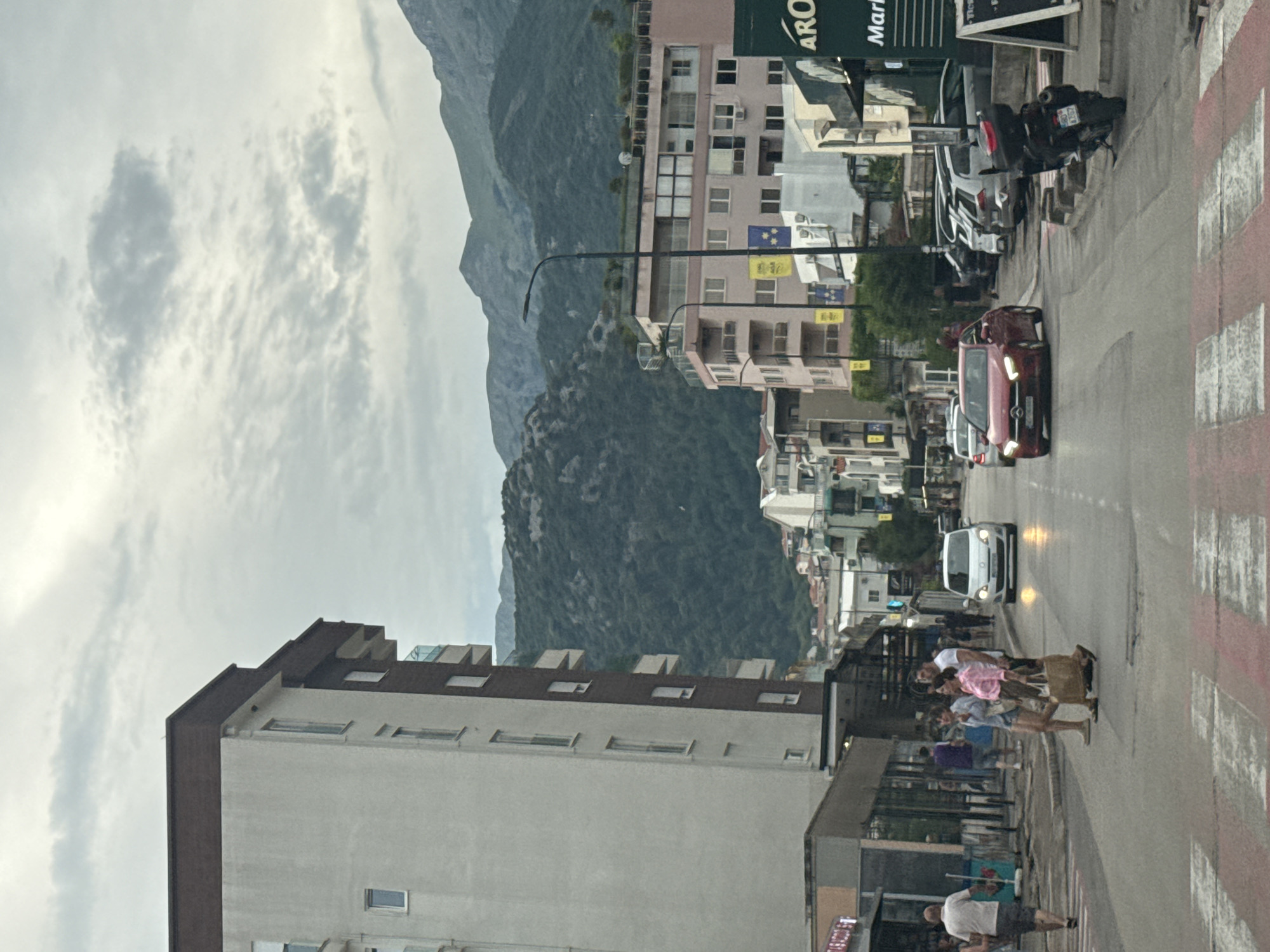 The image captures a street scene in Budva, Montenegro, with a backdrop of imposing mountains and a cloudy sky. In the foreground, a pedestrian crossing with red and white markings is visible. Several people are seen crossing the street or walking on the sidewalk, some appearing to be tourists, judging by their attire and relaxed demeanor. One group of individuals is captured mid-stride on the crosswalk.
Vehicles are present on the street, including a prominent pink car with its headlights on, and a white car further down the road. A motorcycle is parked on the sidewalk. The architecture of the buildings lining the street suggests a Mediterranean coastal town, with multiple stories and balconies. Some buildings have businesses on the ground floor, indicated by signage.
The time of day appears to be late afternoon or early evening, given the lighting conditions and the overcast sky. The weather seems mild, with the clouds suggesting the possibility of rain, but no precipitation is actively visible. The overall mood is that of a bustling yet tranquil urban environment, with the natural beauty of the surrounding landscape providing a striking contrast to the man-made structures.
Visible text includes "ARO" and "Mark" on a sign above one of the businesses. License plates are also visible on some vehicles, though not clearly legible.