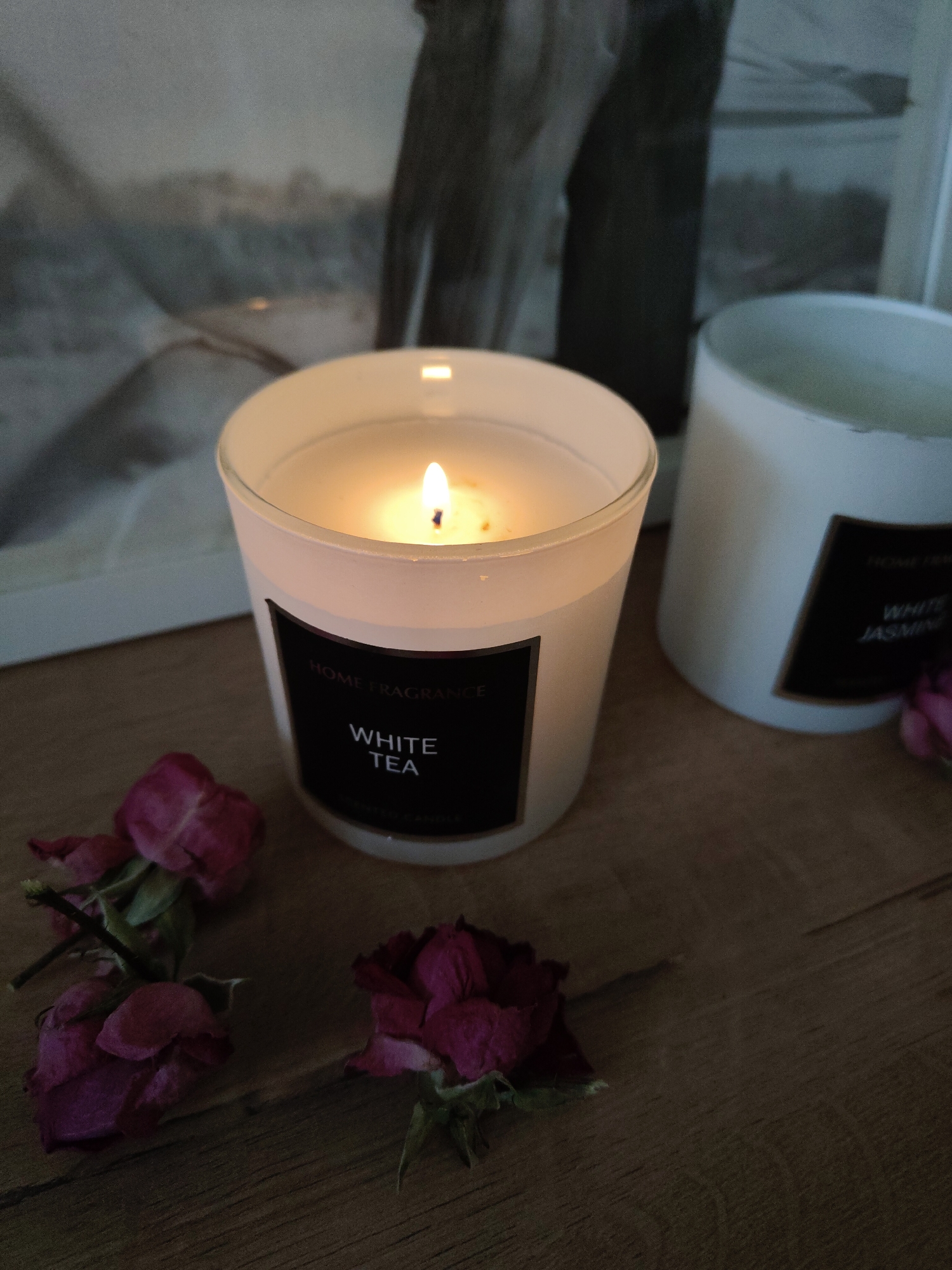 The image shows two candles on a wooden surface, with dried rose petals scattered around them. The candle in the foreground is white with a black label that reads "HOME FRAGRANCE" above "WHITE TEA". It is lit, and the flame is visible. Behind it, slightly out of focus, is another white candle with a black label that appears to read "WHITE JASMINE". A framed picture or artwork is also partially visible in the background. The scene appears to be indoors and well-lit, possibly by the candlelight itself and ambient light. The city and specific location are unknown.
