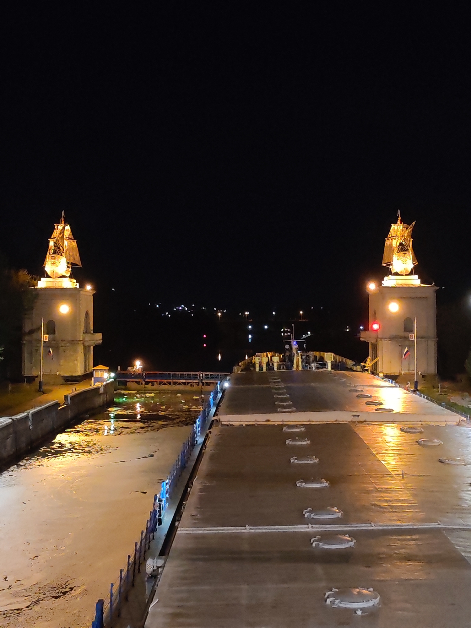Captured at night, the image features the locks of the Moscow Canal in the city of Yakhroma, Russia. Two towers, each topped with an illuminated ship sculpture, stand on either side of a waterway. The structures and canal are brightly lit, contrasting with the dark sky above. Reflections from the artificial light sources create patterns on the water's surface. The water is muddy looking and reflects light. A pedestrian walkway with multiple people is visible in the distance, illuminated by streetlights. The surface of the lock appears wet and reflective, punctuated by multiple round access points. The scene suggests a functional maritime infrastructure with decorative elements.
