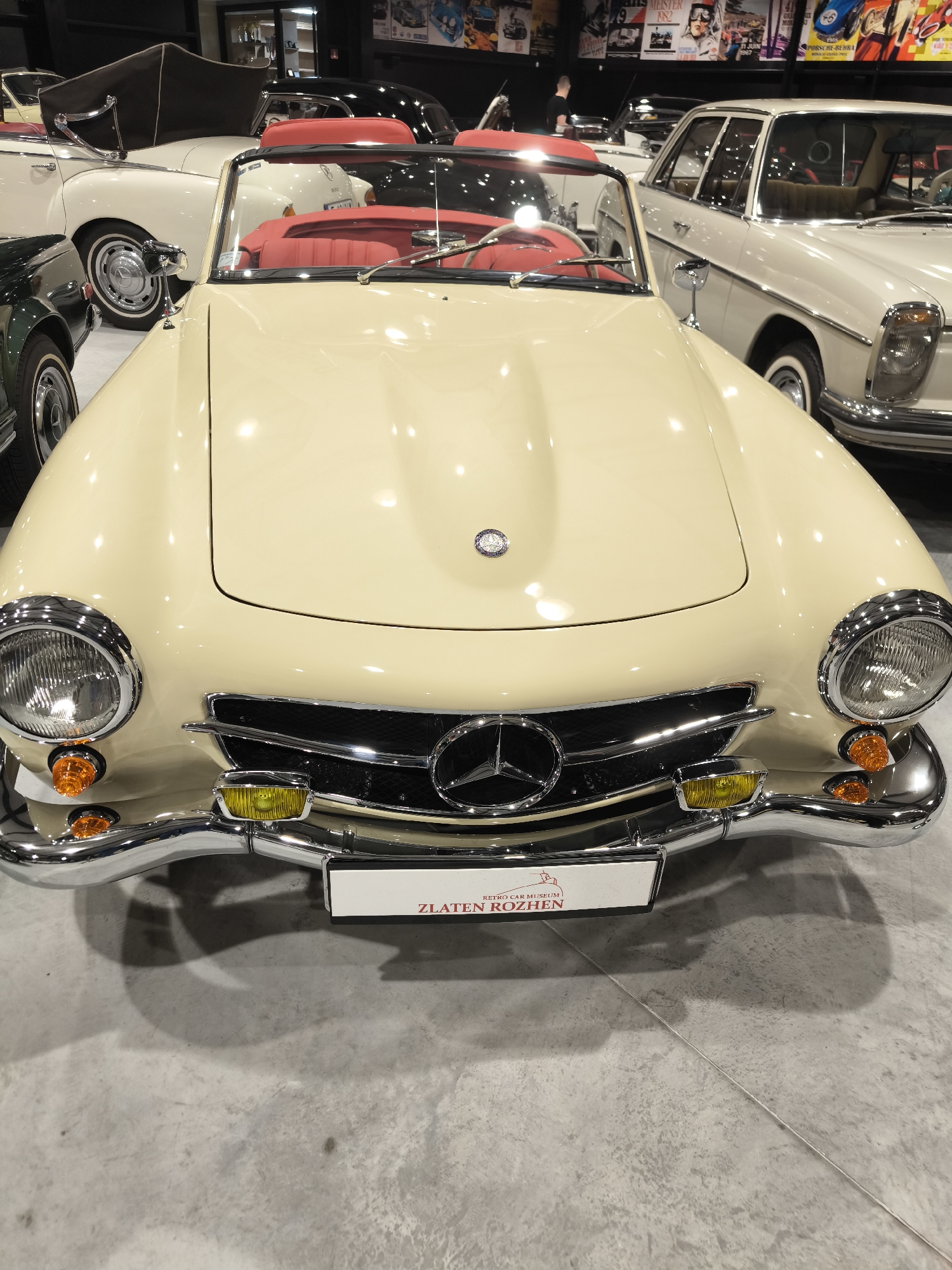 A cream-colored classic Mercedes-Benz 190 SL convertible is parked in what appears to be the Retro Car Museum Zlaten Rozhen in Kapatovo, Bulgaria. The car has a red interior and is surrounded by other vintage vehicles, including a white Mercedes-Benz and a green car. The front of the convertible features a prominent Mercedes-Benz emblem on the grille and hood. The background shows posters or images on the wall, possibly related to cars or the museum. The floor is concrete, and the overall setting is indoors.