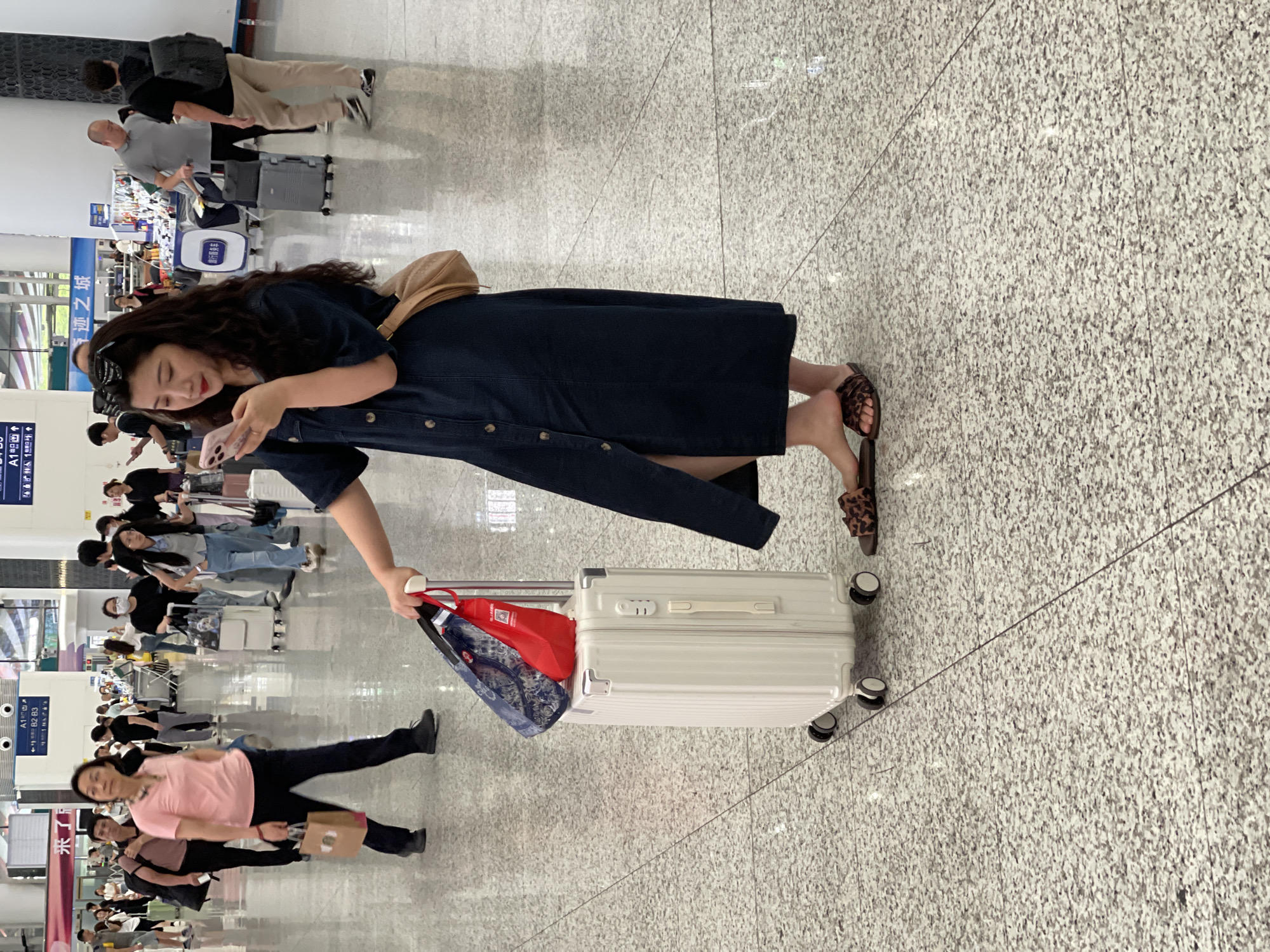 In a well-lit indoor public space, likely an airport or train station in Longhua District, China, a young woman with long, dark, wavy hair stands holding a white suitcase and a pink smartphone. She is wearing a dark denim dress and leopard-print slide sandals. Her phone is positioned in her right hand, and she appears to be looking at its screen. Two bags, one blue with a patterned design and one red, are attached to the handle of the suitcase. The suitcase has four wheels and a silver handle. In the background, numerous other people are visible, some walking with luggage, others standing. The floor is made of speckled gray tile. Signs in Chinese characters are visible on the walls in the distance.