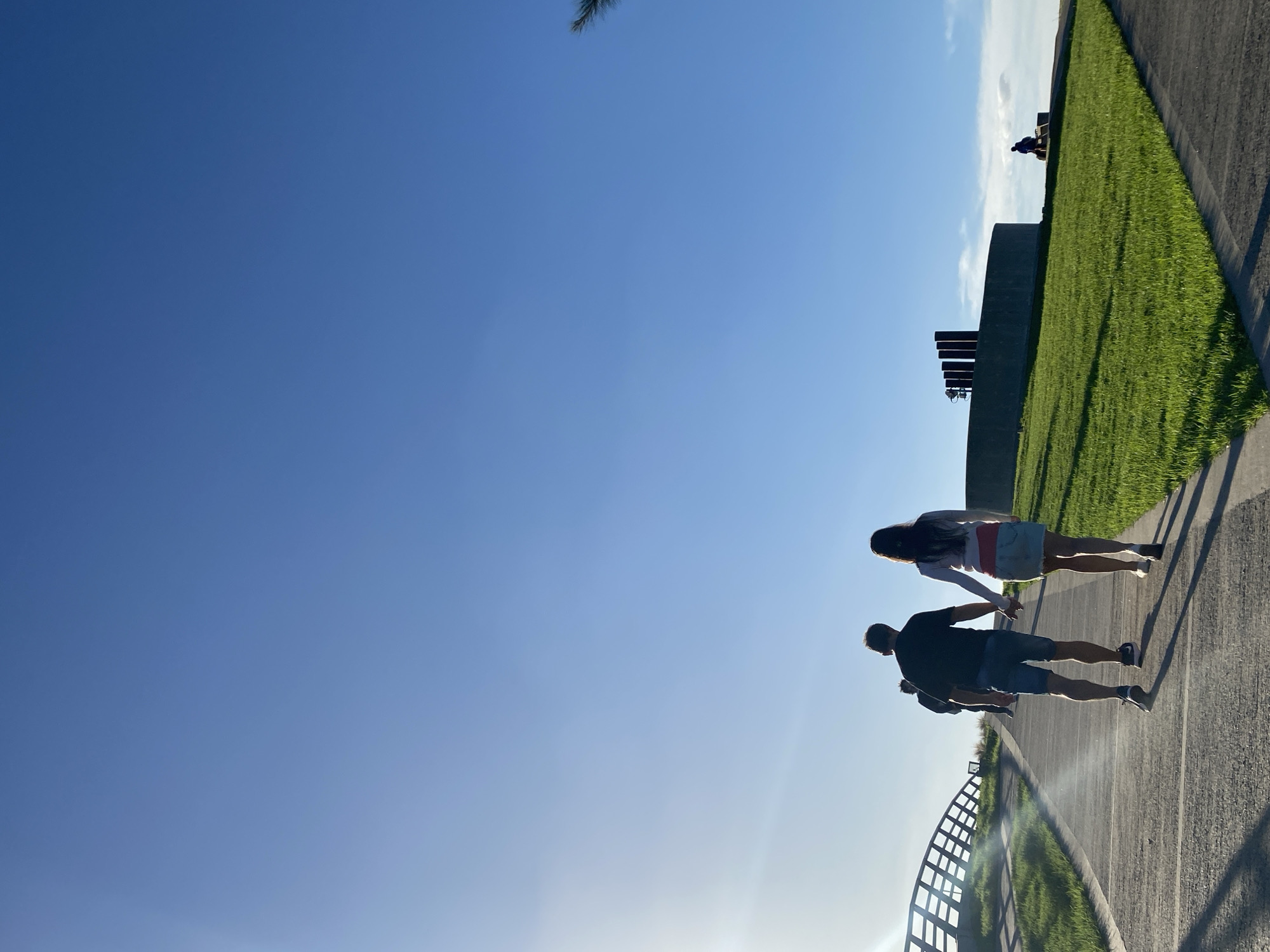 A low-angle shot shows a couple walking hand-in-hand along a waterfront promenade in Buenos Aires, Argentina. The camera angle suggests the image was taken from a low position, near the railing. The couple is walking away from the camera. The promenade is made of concrete, and a metal railing borders the water. To the right of the promenade is a grassy area with a low wall and benches overlooking the water. The sky is a clear blue, with some glare from the sun. The location is likely Puerto Madero.