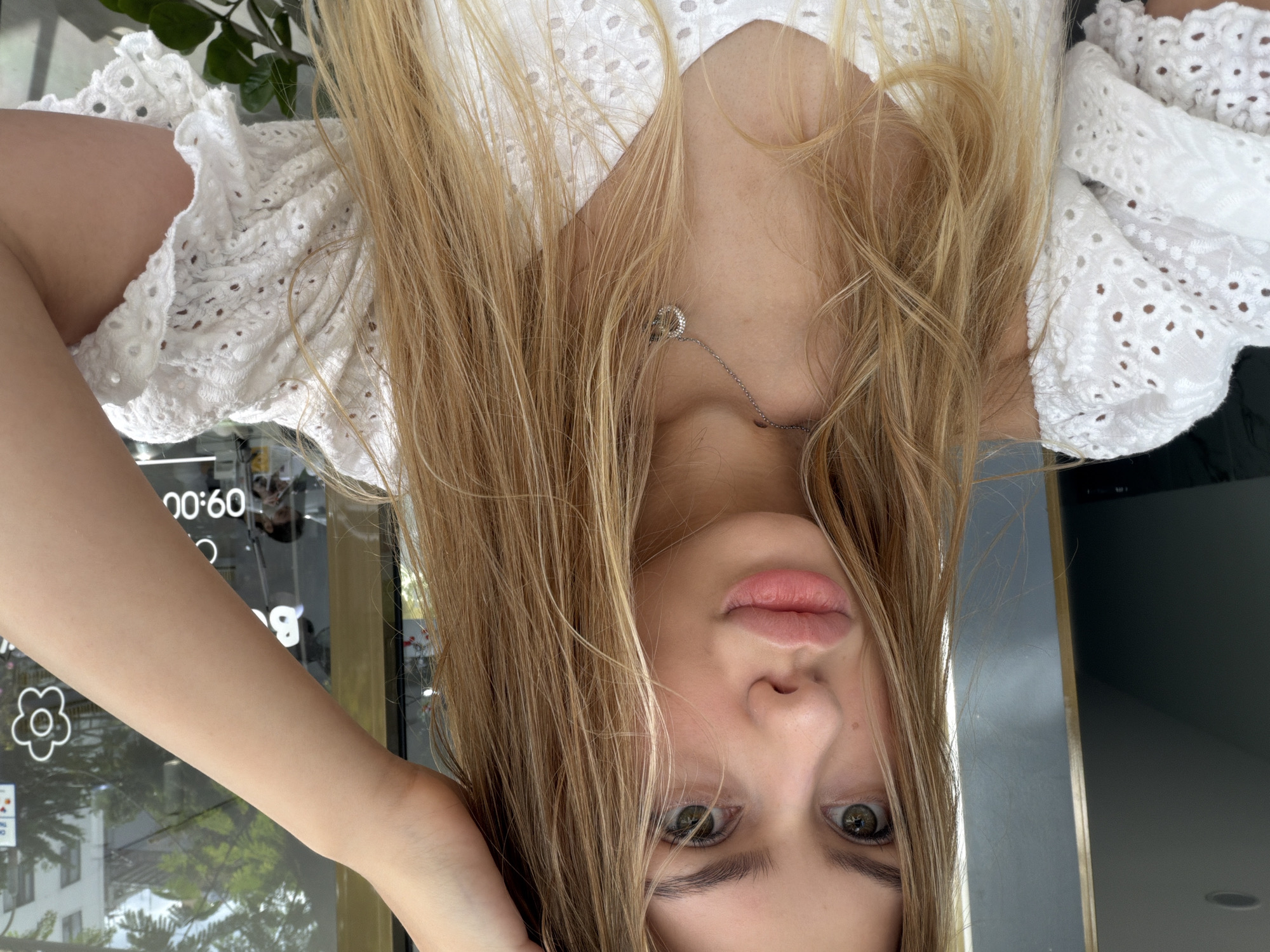 A young woman with long blonde hair is posing for a selfie in front of a store window with the time 09:00 visible on a sign. She is wearing a white eyelet blouse and a silver necklace. Her left hand is raised to her head, and her right arm is visible. The background shows buildings and greenery, indicating an outdoor urban setting. The image is taken in Đà Nẵng, Vietnam.