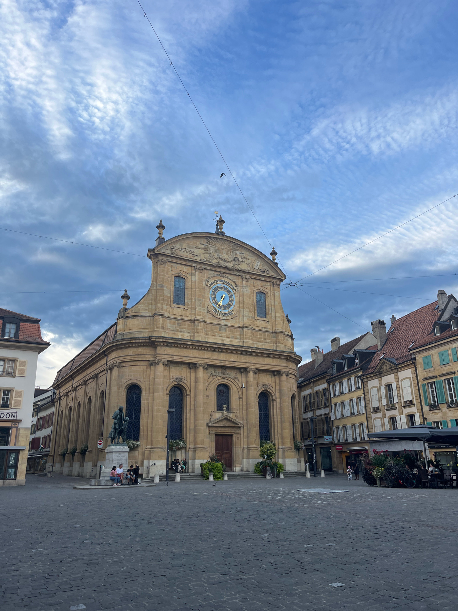 The image depicts the Place Pestalozzi in Yverdon-les-Bains, Switzerland, featuring the historic Temple de Yverdon in the center. The Temple is a large, light-brown stone building with a classical facade, a prominent clock face with Roman numerals above the main entrance, and arched windows. To the left of the Temple, a statue of a man and two children stands on a pedestal. Several people are seated on benches around the statue and on the steps of the Temple. The square itself is paved with dark cobblestones. To the right of the Temple, a row of multi-story buildings with traditional European architecture lines the square, some featuring awnings and outdoor seating for cafes. The sky above is partly cloudy, with a mix of blue sky and white, wispy clouds, and a few wires are visible crisscrossing the sky. The lighting suggests it is late afternoon or early evening.