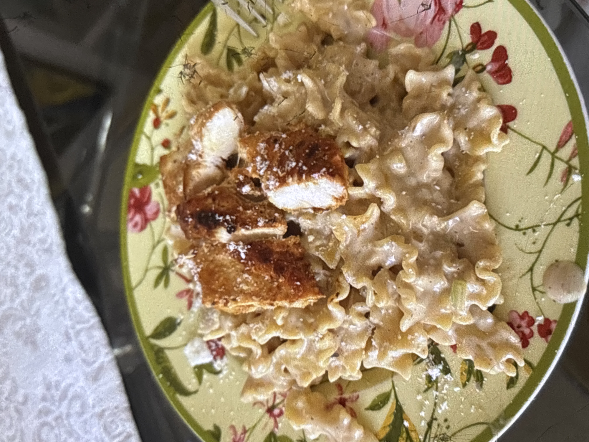 A close-up, overhead shot shows a serving of campanelle pasta with a creamy sauce and pieces of grilled chicken breast on a decorative plate. The plate is pale yellow with a green rim and features a floral pattern of red flowers and green leaves. The pasta is generously coated in a light-colored sauce, and sprinkled with grated cheese. The chicken pieces are golden brown and appear to be seasoned. The background is blurred, but suggests a textured surface, possibly a tablecloth, and a dark, reflective surface, likely a table. There is no indication of the city of Orihuela, Spain.
