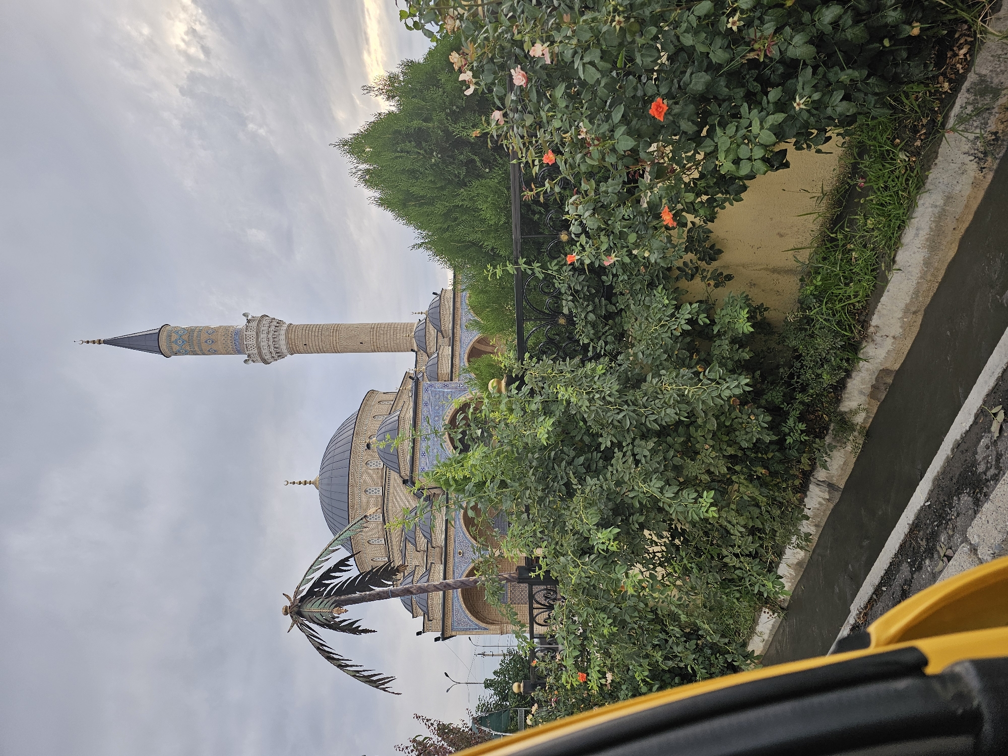The image captures a mosque with a tall, slender minaret and a large dome, characteristic of Islamic architecture, situated in the city of Özgön, Kyrgyzstan. The mosque is adorned with intricate blue tilework on its exterior. In the foreground, lush green bushes and flowering rose bushes, some bearing orange and pink blossoms, are visible, separated from the mosque by a black metal fence. A decorative palm tree stands to the left of the mosque. A yellow object, possibly part of a vehicle, is in the lower left corner, partially obscuring the view. A narrow concrete drainage channel runs along the roadside in front of the vegetation. The sky is overcast with clouds, and the lighting suggests it might be early morning or late afternoon.