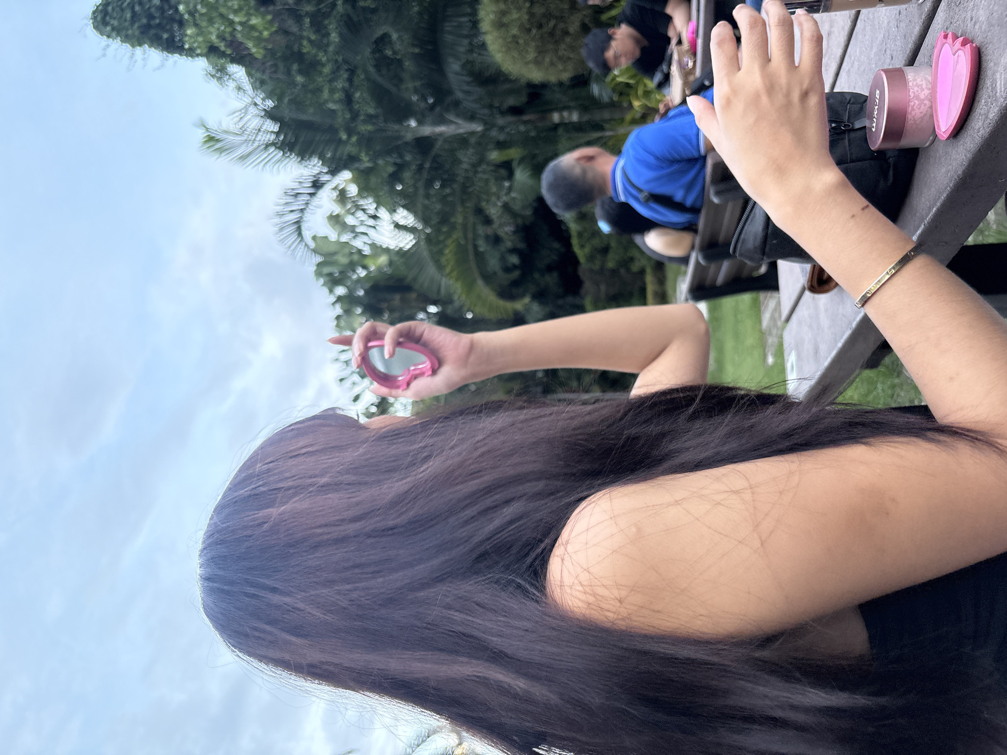 The image shows a woman with long black hair sitting at a gray picnic table in an outdoor setting. She is holding a small pink handheld mirror in her left hand. On the table next to her right hand, which has a gold bracelet, are a makeup compact and a container of makeup. In the background, partially obscured by greenery, are two men sitting at the same table. The location is Davao City, Philippines. The sky is overcast.