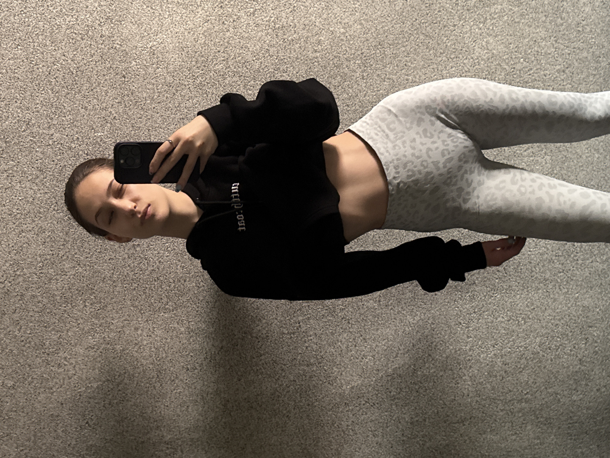 A young woman is lying on a carpeted floor taking a selfie. She is wearing a black, cropped hooded sweatshirt and light gray leggings with a leopard print pattern. The woman is holding a black smartphone. The background is a plain, gray carpet. The location is likely indoors. There is no information to indicate the city of Sofia, Bulgaria.