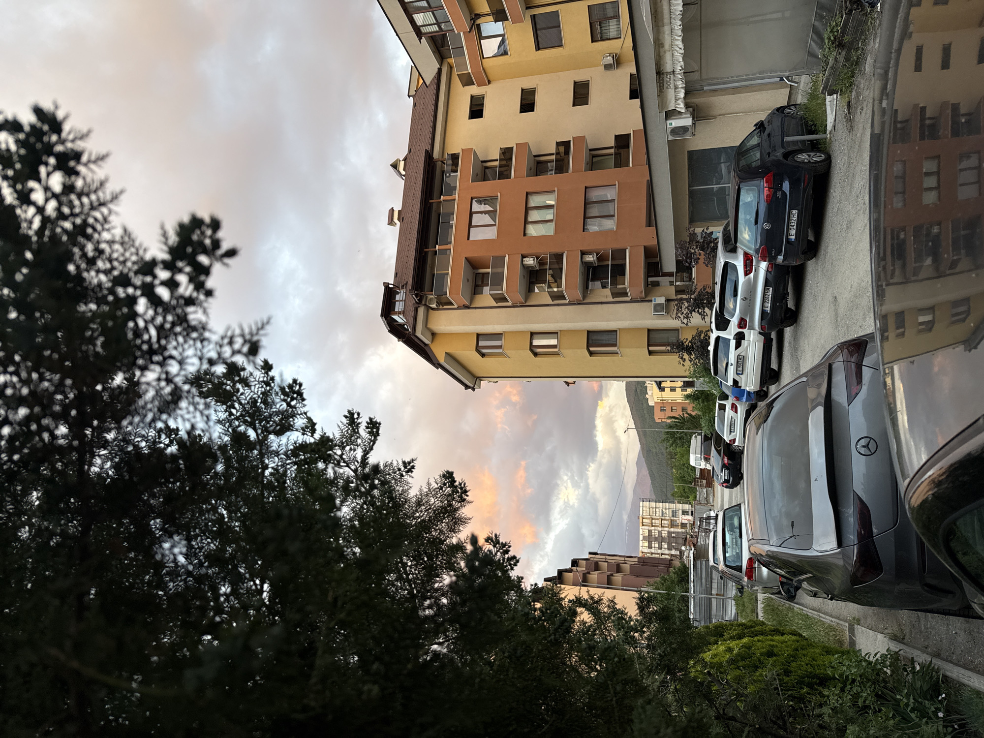 The image is a ground-level shot of a street scene in what appears to be a residential area of Blagoevgrad, Bulgaria, judging by the license plates on the vehicles. Several cars are parked along the side of the road, including a dark blue Volkswagen Golf (license plate E 0432 HC), a white Renault Kadjar (license plate E 2262 HM) and a gray Mercedes-Benz sedan partially visible in the foreground. The cars are parked close together on a street next to the sidewalk. The background features multi-story apartment buildings with beige and brown exteriors. Between the buildings and above the horizon, the sky shows clouds with hints of orange from the setting sun. To the left of the buildings, there are trees with dark green foliage. The foreground includes bushes. The road surface appears to be asphalt and the image appears to have been taken during twilight.
