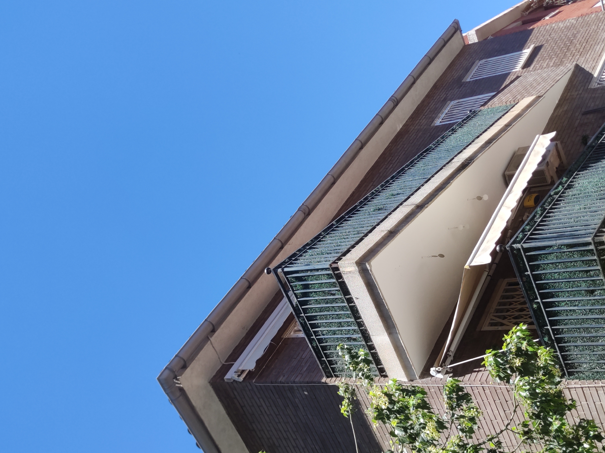 A low-angle, tilted shot captures a building under a clear blue sky. The building, constructed of dark brown brick, features protruding balconies with dark green railings. A white awning is extended from one of the balconies. The building also features rectangular vents on the facade. Some green foliage is visible near the bottom of the frame. The scene appears to be from an urban location.