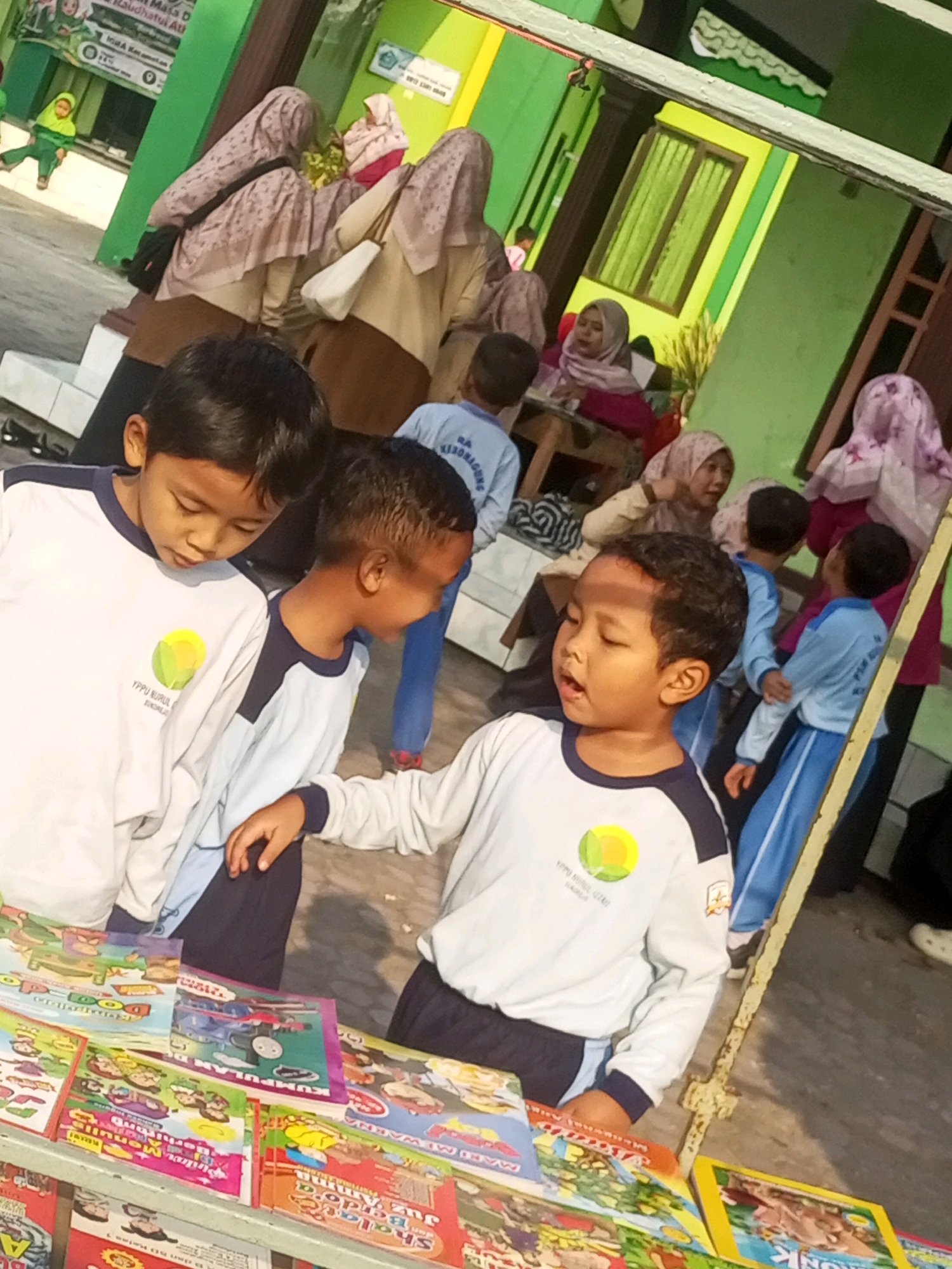 In the city of Gurah, Indonesia, this image captures an outdoor scene likely at a school. Two young boys in white and navy blue school uniforms are the central focus. The boy on the left is looking down, while the boy on the right is looking forward with his mouth open. In the background, several other children and adults are visible, some wearing headscarves. Some of the children are wearing similar school uniforms. In the foreground, a display of colorful children's books is laid out on a shelf or table. The environment appears to be a sunny day.