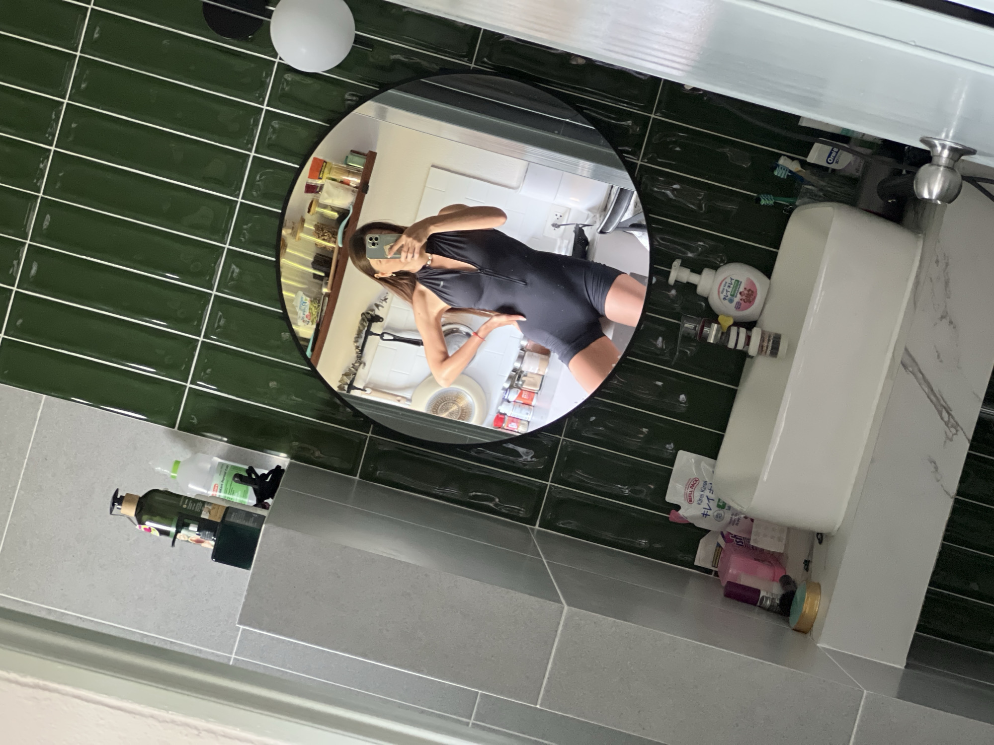 Here is a forensic description of the image:
**Scene:** The image captures a bathroom interior, primarily focusing on a mirror reflection.
**Environment:** The bathroom features green, horizontally-laid, subway tiles on one wall and gray tiles on a portion of another. There is a white sink and vanity, and a white cabinet above the sink. The lighting is generally bright.
**Subjects:**
* A person is visible in the mirror reflection. The individual is holding a mobile phone up, ostensibly taking a selfie. The person is wearing a black, form-fitting, halter-style bodysuit.
**Objects:**
* Mirror: The round mirror is the central focal point, reflecting the bathroom's contents and the person taking the picture.
* Sink: A white sink sits near the mirror, positioned towards the right side of the frame.
* Toiletries: Bottles of soap, skin lotion, toothpaste, and other toiletries are placed on the vanity top.
* Lighting Fixture: A circular light fixture, with a white glass globe, is installed on the tiled wall.
* Mobile Phone: The person is holding a mobile phone, presumably to take a photo.
**Location:** Unknown, Unknown.