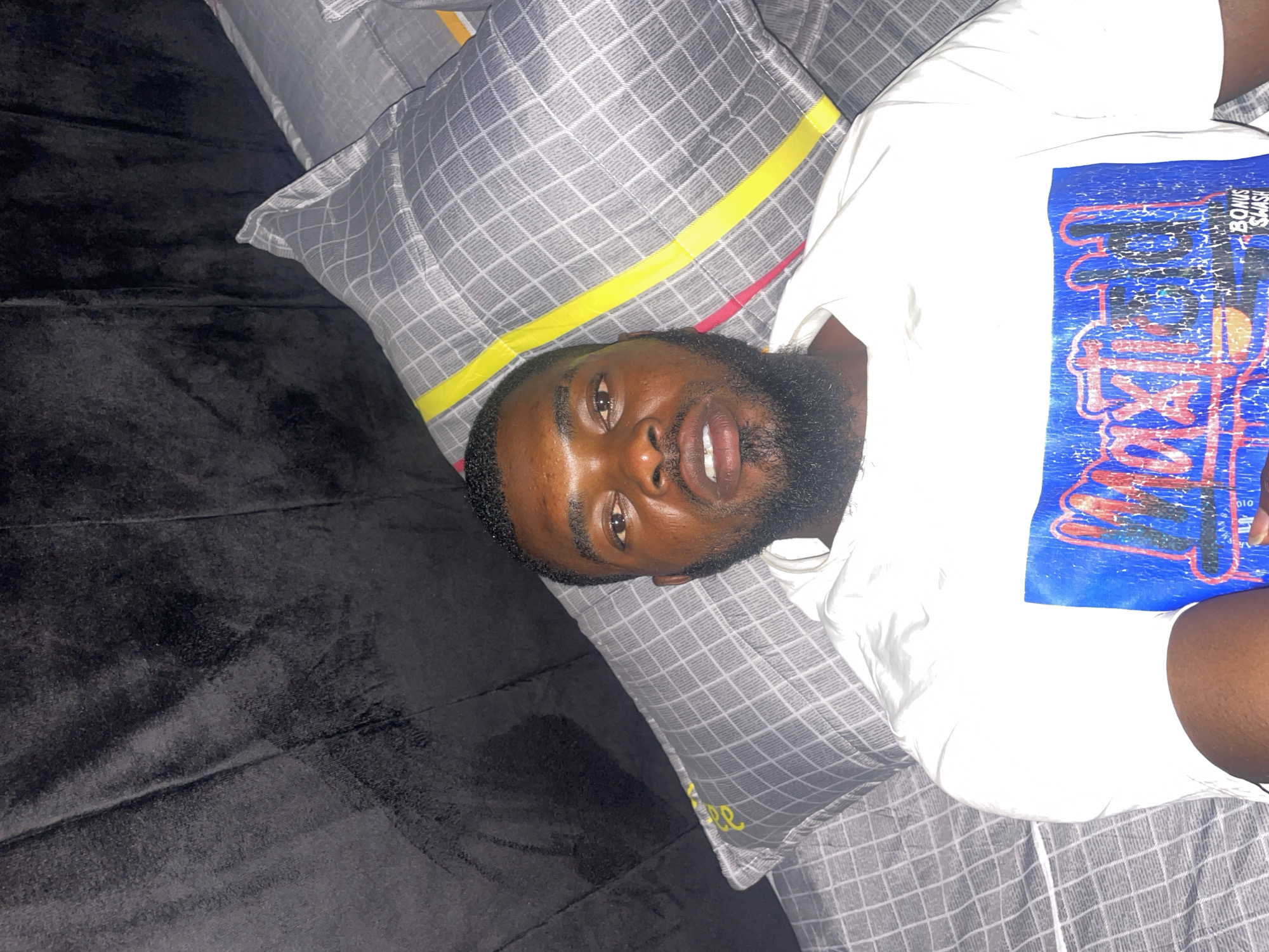 A man with dark skin and a beard is lying down on a bed. He is wearing a white T-shirt with a blue graphic. The bed has a gray plaid pillow with yellow accents and a black blanket. The location is Jos, Nigeria.