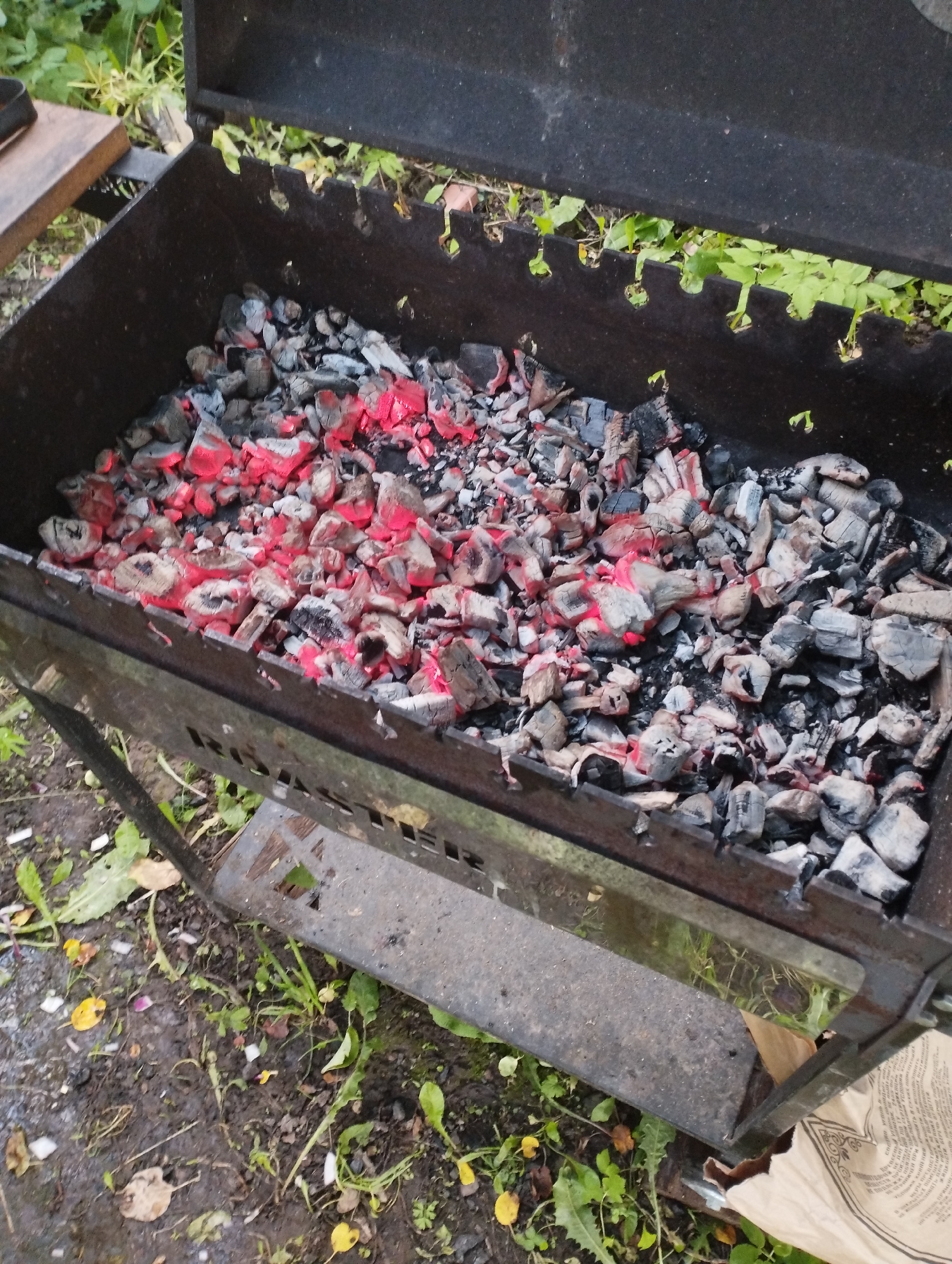 A close-up, high-angle shot shows a metal barbecue grill filled with glowing red and grey charcoal, situated outdoors on a patch of dirt and greenery, possibly in the city of Ширяево (ильинский), Russia. The grill itself is a dark metal rectangle with a hinged lid, the front of which is slightly ajar, revealing the hot coals within. The serrated edge of the grill's interior is visible, designed to hold cooking grates. The ground around the grill is covered in wet, dark soil, scattered with fallen leaves and small green plants. A piece of wooden material is visible to the upper left, and a crumpled paper bag with text is partially seen at the bottom right. The lighting suggests it is daytime, with some overcast conditions. There are no people or animals present in the image.