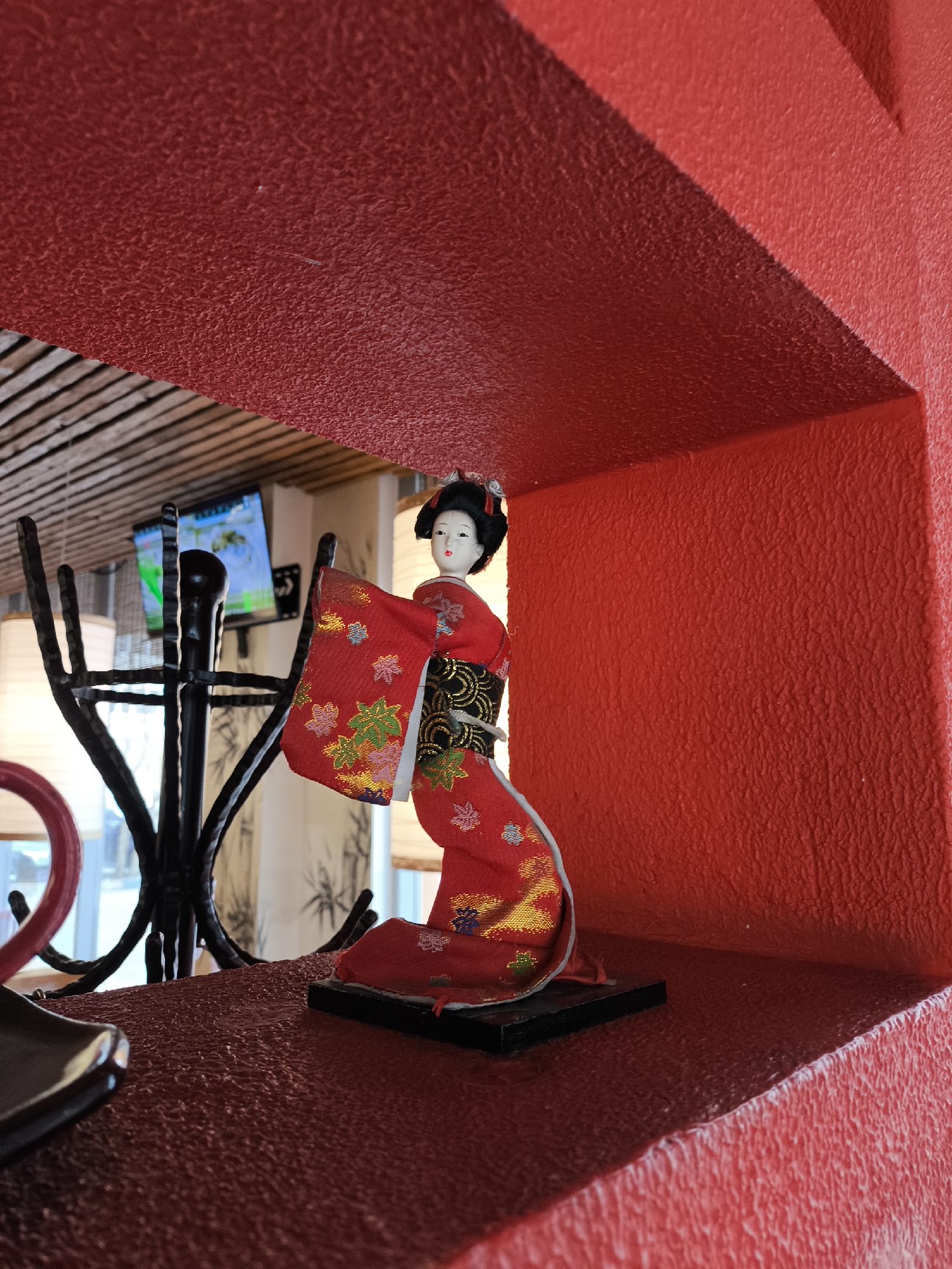 The image shows a Japanese doll displayed in a red alcove. The doll wears a traditional red kimono with floral patterns. The surrounding environment includes decorative ironwork and a glimpse of a window and a television. There is no information to identify the location of the image in the city of Moscow, Russia.