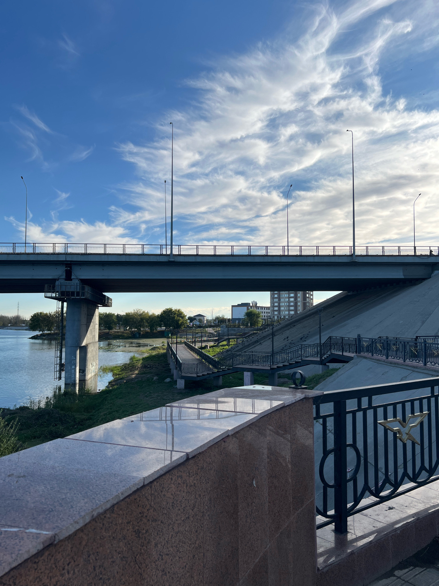 The image captures a wide shot of a riverfront promenade and a bridge in Kostanay, Kazakhstan. In the foreground, a polished granite wall and a black metal railing with an ornate bird emblem line the promenade. Beyond the promenade, a concrete embankment slopes down to the river. A wooden boardwalk with metal railings winds its way along the riverbank. The river itself is calm, reflecting the blue sky and wispy white clouds.
Dominating the mid-ground is a large concrete bridge spanning the river, supported by a massive concrete pillar. The bridge deck is lined with railings and streetlights. In the distance, several multi-story buildings are visible, including a taller, modern building. Trees dot the landscape along the riverbanks. A single figure is visible walking on the bridge. The sky is a vibrant blue with scattered, feathery clouds, suggesting a clear day with a gentle breeze. The overall impression is one of a well-maintained urban public space with a natural element.