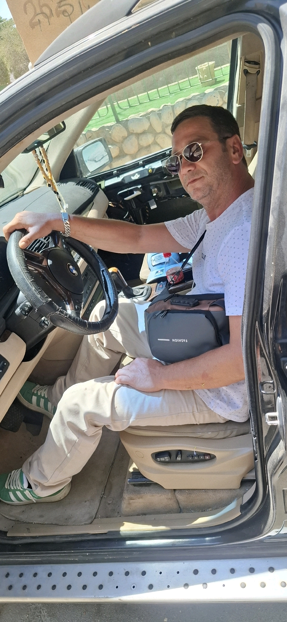 A man is sitting in the driver's seat of a car, holding the steering wheel. He is wearing sunglasses, a white t-shirt with a subtle pattern, and light-colored pants. He also has a small dark bag across his chest. His left hand is on the steering wheel, and his right arm is resting on his leg. The interior of the car is visible, including the dashboard and the seats. Outside the car, a rocky wall and some greenery can be seen. A sign with some writing is visible on the car's roof. The scene appears to be in Dimona, Israel, suggested by the sign's markings that resemble Hebrew characters and the general environment.