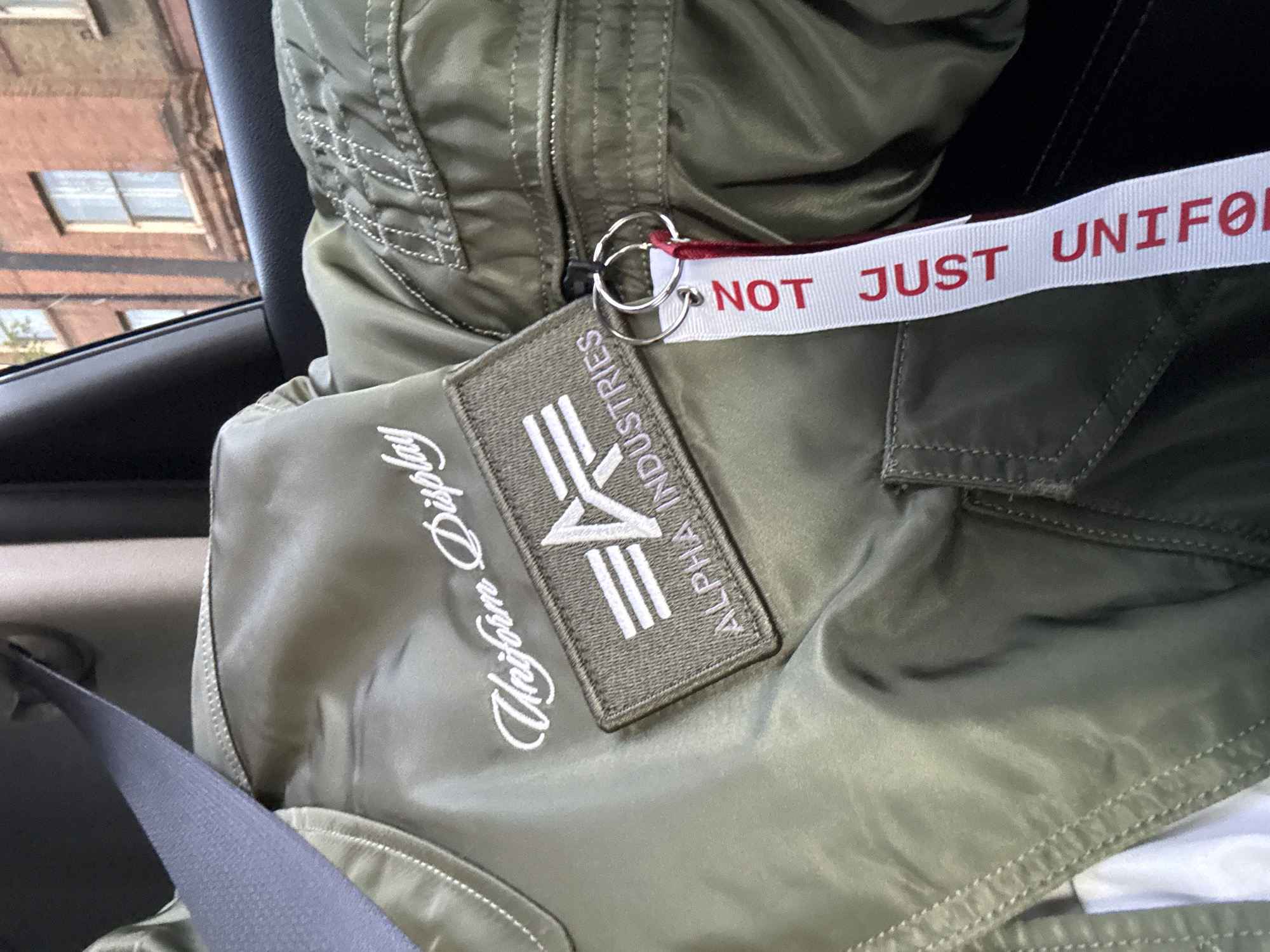 The image shows an Alpha Industries bomber jacket, likely indoors in a vehicle. The jacket is olive green and features a patch with the Alpha Industries logo and "Uniform Display" stitched in white cursive. A white lanyard with the words "NOT JUST UNIFOR" printed in red is attached to the jacket's zipper. A seatbelt crosses over the jacket. Through the car window, a building with windows is partially visible, suggesting an urban environment. No specific London landmarks are discernible to pinpoint the location.