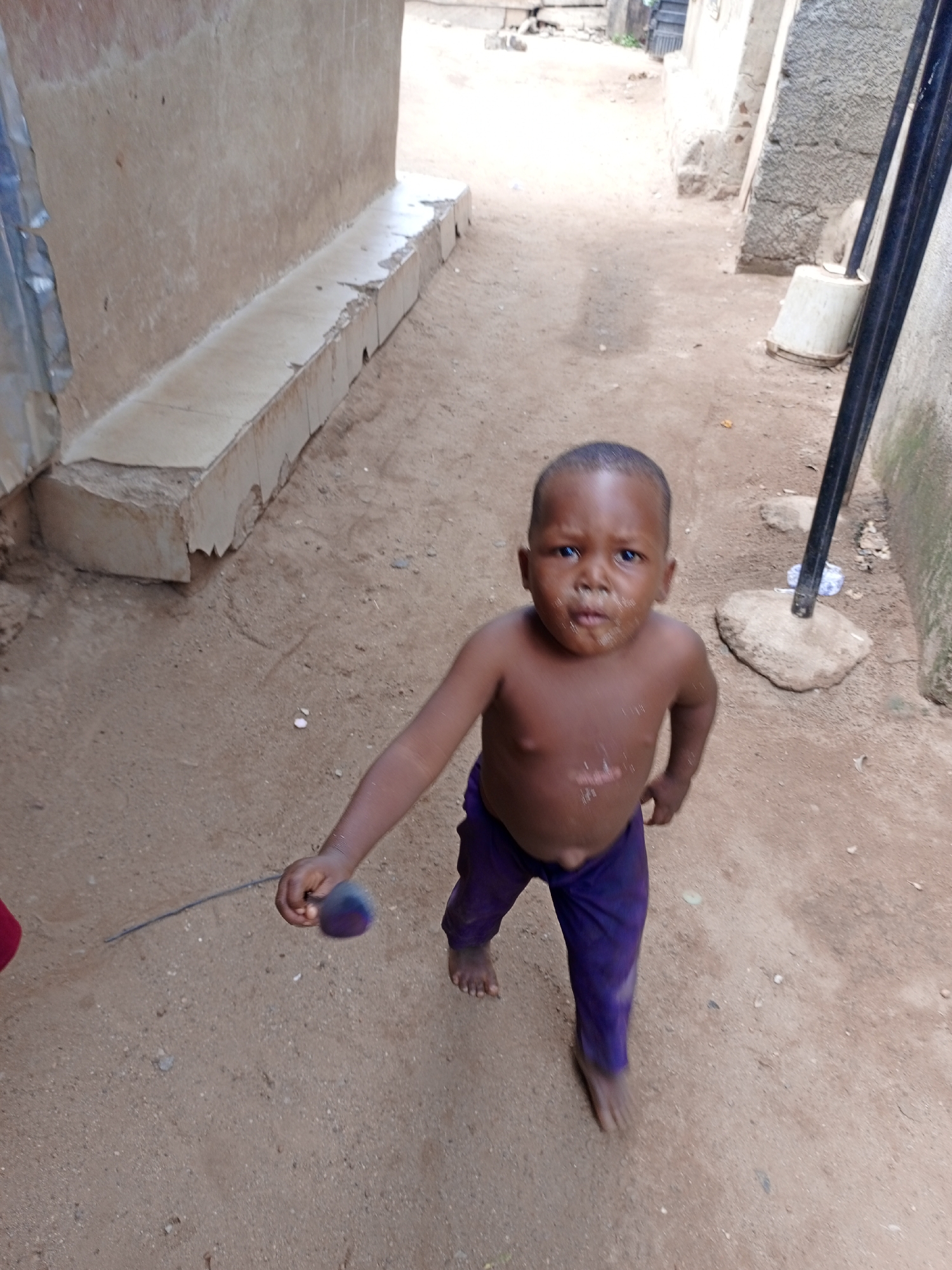 A young child stands barefoot on sandy ground in what appears to be an alley or narrow pathway in Keffi, Nigeria. The child, with a dark complexion and a slightly dirty face, wears purple pants and no shirt. The child holds a small, dark object attached to a thin, dark wire or string in their right hand.
The background shows buildings with concrete and stone walls. To the left is a low concrete ledge or bench. Further back, the alley opens up slightly, showing more buildings and some vegetation. A metal pole is visible on the right side of the frame. The ground is a mix of sand and dirt, with scattered debris.