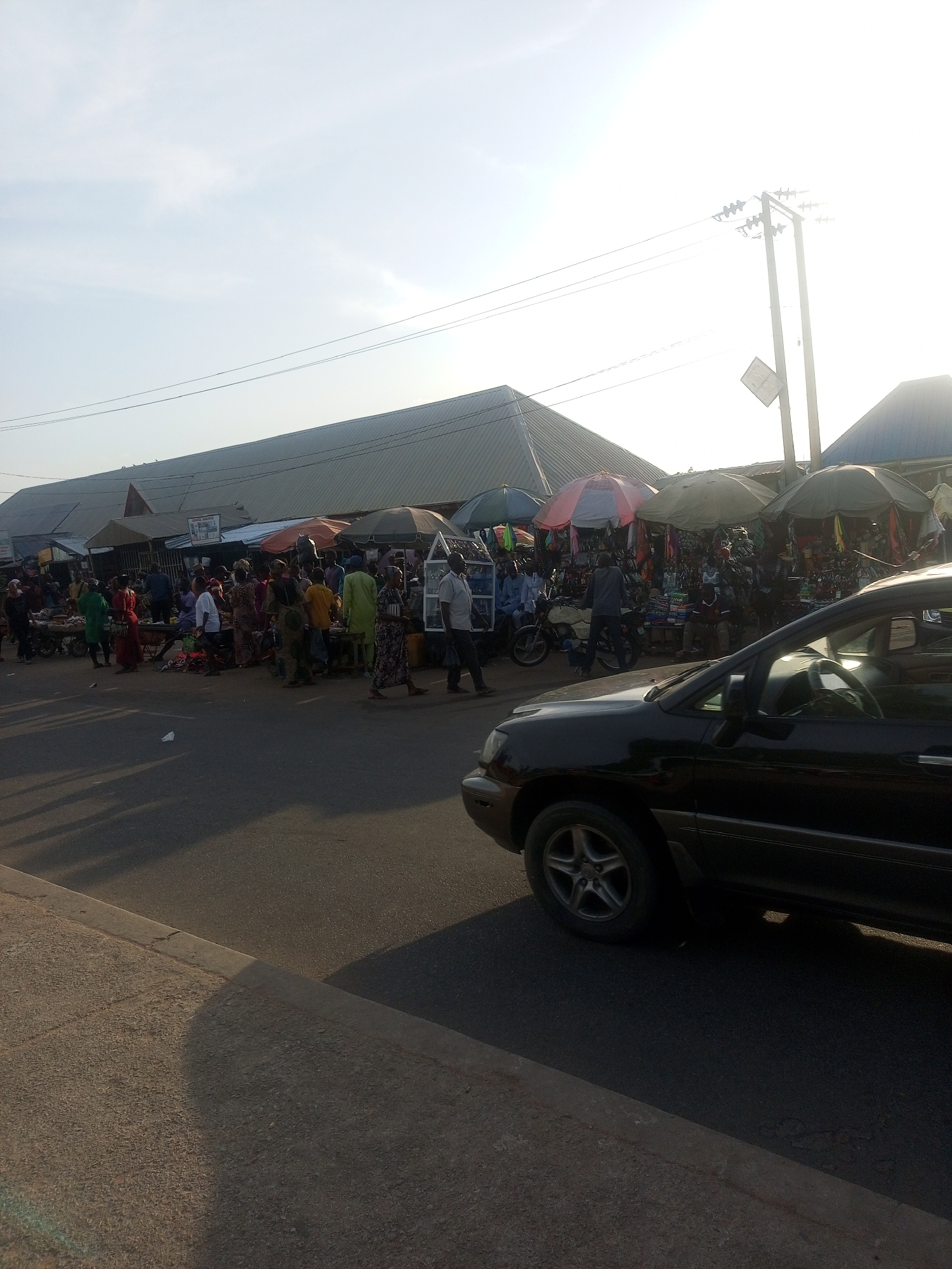 The image depicts a bustling outdoor market scene in Jalingo, Nigeria. A busy street runs along the foreground, with a black SUV partially visible on the right. The market itself is set up along the street, with numerous stalls covered by umbrellas and awnings. A variety of goods are displayed, including clothing, textiles, and what appear to be household items.
Many people are actively engaged in the market. They are seen browsing, interacting with vendors, and moving between stalls. Some individuals are dressed in traditional African attire, while others wear more contemporary clothing. The overall atmosphere suggests a lively and active commercial environment.
In the background, a large, corrugated metal structure with a sloped roof dominates the skyline, likely a permanent market building or warehouse. Power lines and a utility pole with a diamond-shaped sign are also visible against the bright, hazy sky. The sunlight suggests it is daytime, possibly late afternoon given the long shadows cast on the road. The weather appears to be clear but hazy, possibly due to dust or pollution common in the region. No specific emotions are discernible from the crowd, but the general activity indicates a sense of purpose and commerce. There is a sign visible on one of the market stalls, but the text is too small to be legible.