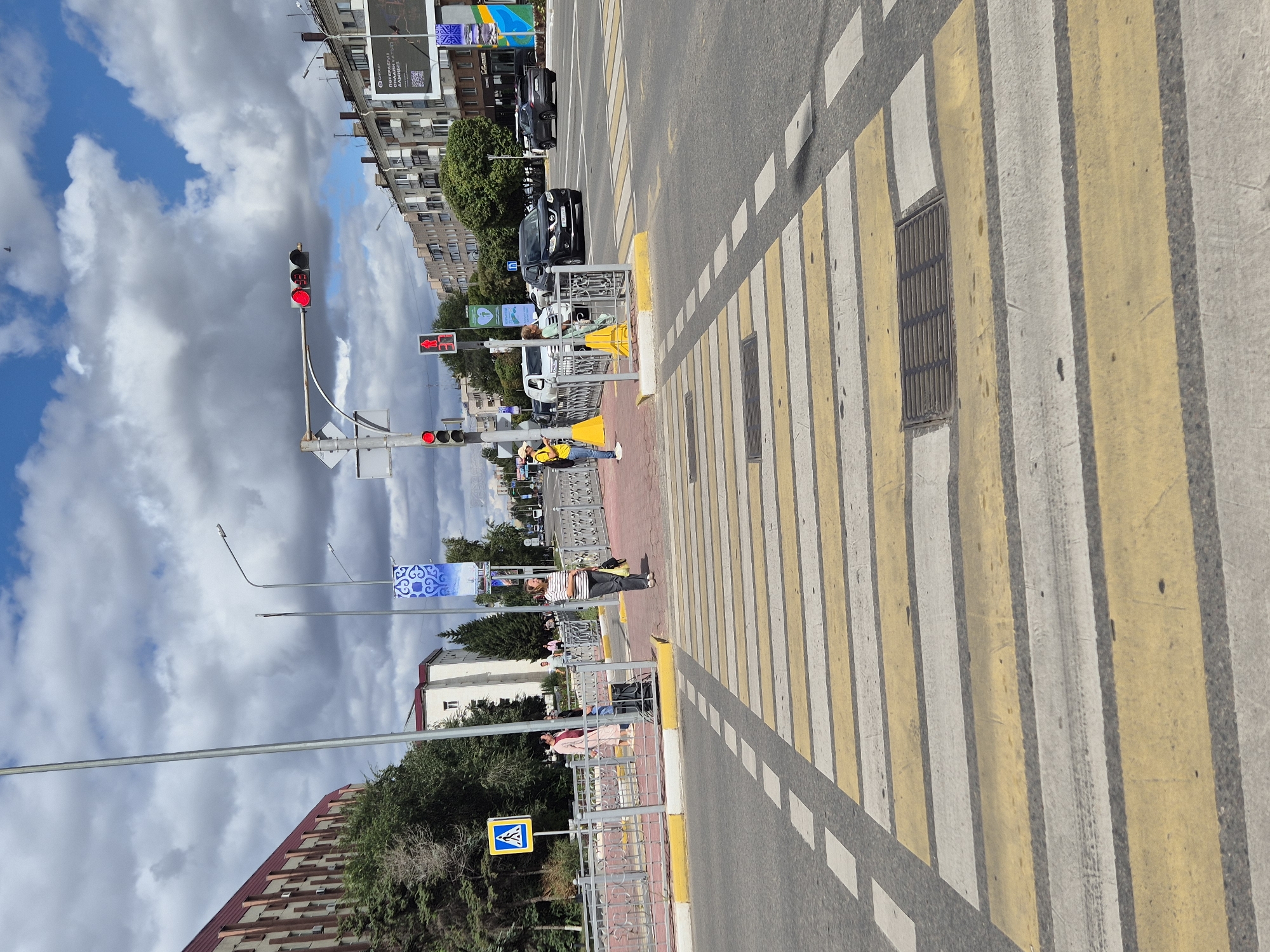An outdoor, eye-level, medium shot captures a pedestrian crossing in Kokshetau, Kazakhstan, on a cloudy day with patches of blue sky. The foreground is dominated by a wide, yellow and white striped pedestrian crossing, with a metal grate embedded in the asphalt near the center. To the left, a pedestrian safety island is marked by a yellow curb and a yellow and blue pedestrian crossing sign. A woman in a pink dress and a woman in a striped shirt and dark pants are standing near this island. A few other pedestrians are visible on the sidewalk behind a metal fence. In the background, a traffic light shows a red signal. Further down the street, cars are driving, and multi-story buildings line the right side of the road. The overall scene depicts a typical urban streetscape with traffic and pedestrian activity.
