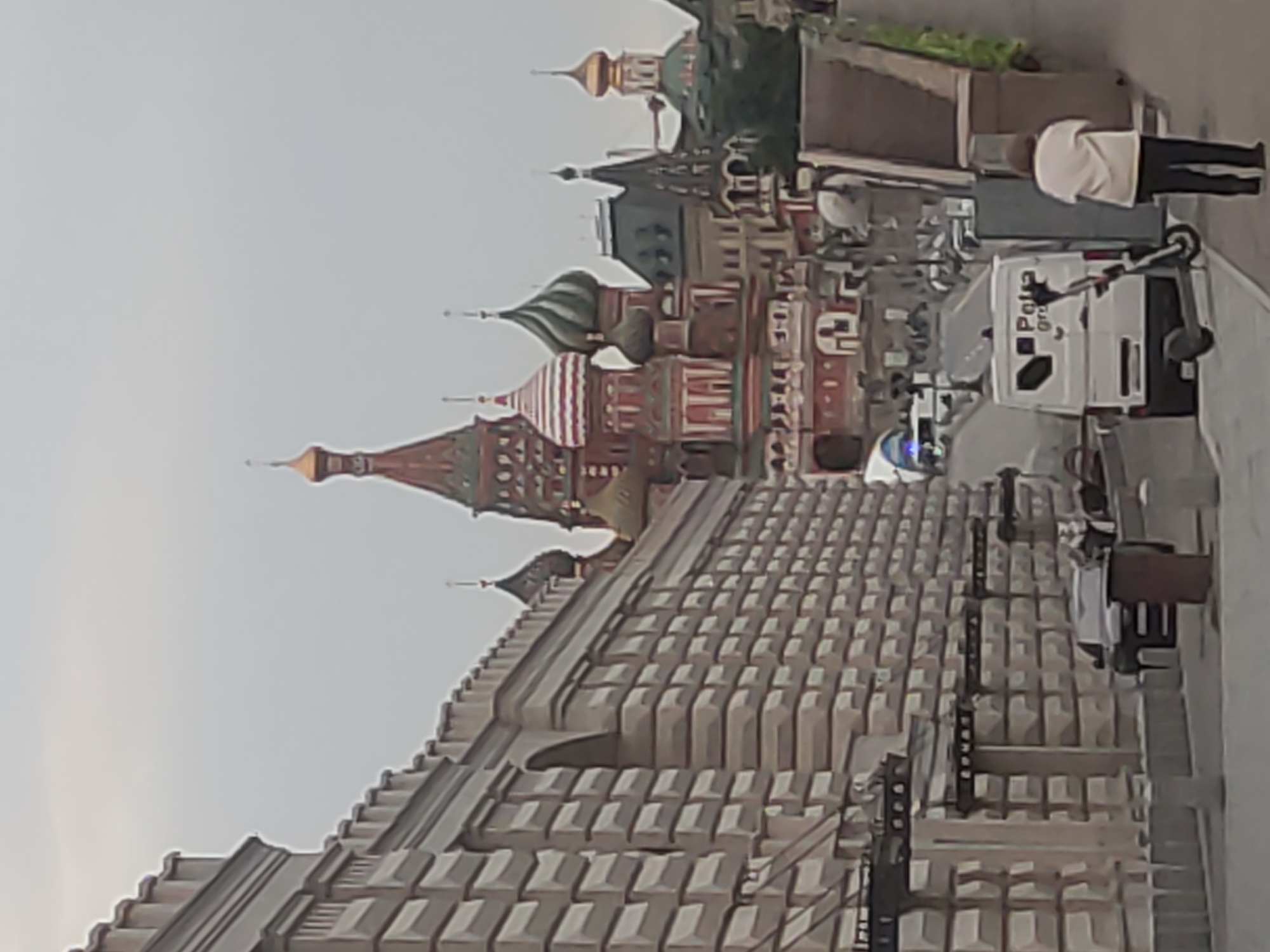 The image shows a partial view of St. Basil's Cathedral in Moscow, Russia, seen from behind the walls of the Kremlin. A white van with the word "Police" on the side is parked on a street, and a person in a white shirt is standing nearby. An electric scooter is also visible. The sky is overcast.