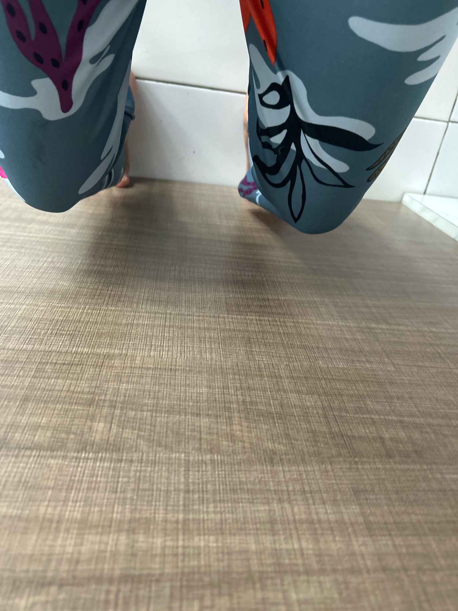 The image shows a low-angle view of a person's legs wearing patterned leggings. The leggings are primarily teal with white, pink, and black floral and leaf designs. The person's feet are bare and visible, one slightly forward of the other. They are positioned against a white tiled wall and a light brown wooden floor. The scene is indoors, in what appears to be a room with a tiled backsplash and wooden flooring. There are no specific landmarks or features that indicate a location in Sukasetia, Indonesia. The focus is on the lower half of the body and the immediate surroundings.