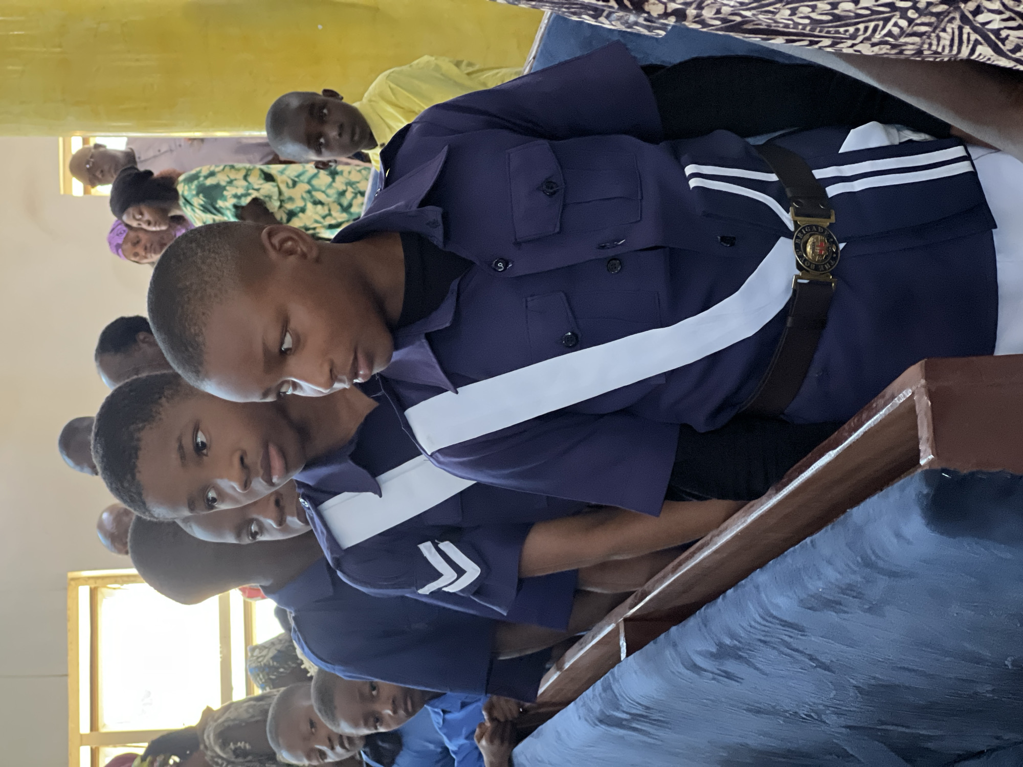 In an indoor setting that appears to be a gathering or ceremony in Jos, Nigeria, several young boys are lined up, some wearing dark blue uniforms with white sashes and belts. The boy in the foreground, on the right, is wearing a dark blue collared shirt with two chest pockets, a white sash crossing his chest, and a black belt with a circular emblem. To his left, another boy in a similar uniform is visible. Behind them, other boys in uniform and civilian attire can be seen, along with adults in the background. One adult is wearing glasses and a light-colored shirt, and another woman is in a green and yellow patterned dress. The background features a bright yellow column and a window with natural light filtering through. The overall atmosphere seems to be one of order and ceremony.