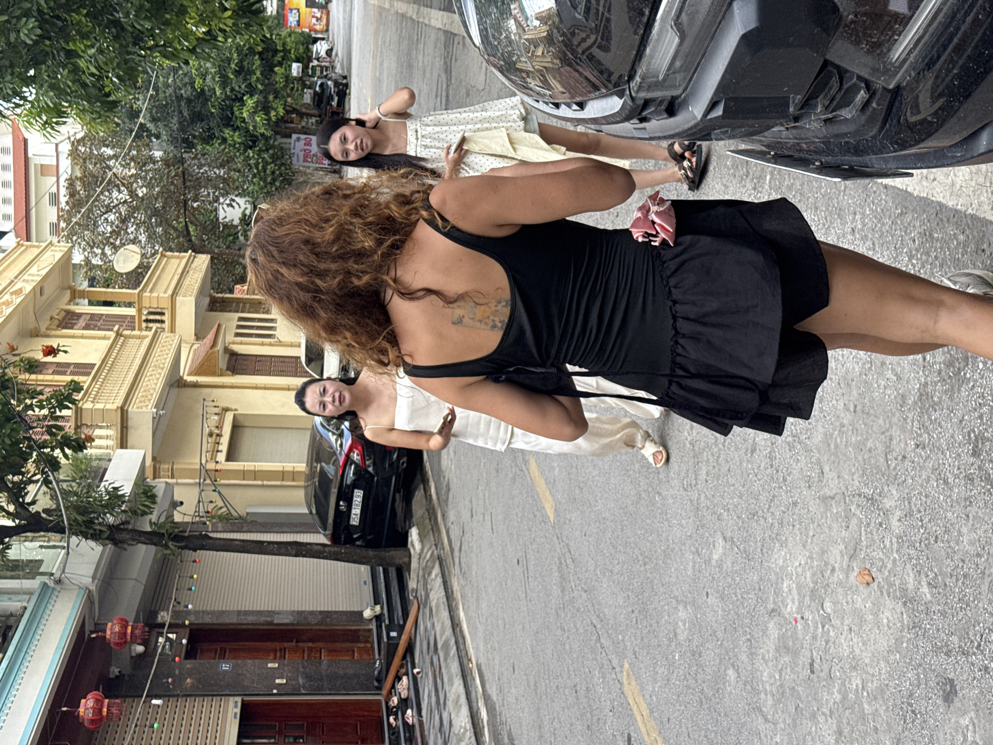 Three women are on a street in Hoa Lư Ward, Vietnam. In the foreground, a woman with curly brown hair and a black dress faces away from the camera. She has a tattoo on her back. To her left and further down the street, a woman in a white sleeveless top and pants holds a phone. To the right of that woman, another woman in a short white dress looks towards the first woman. All three women appear to be walking or standing on the asphalt street, which has a yellow line marking. Buildings with balconies and windows line the street, and trees are visible in the background. A black car is parked on the right side of the image.