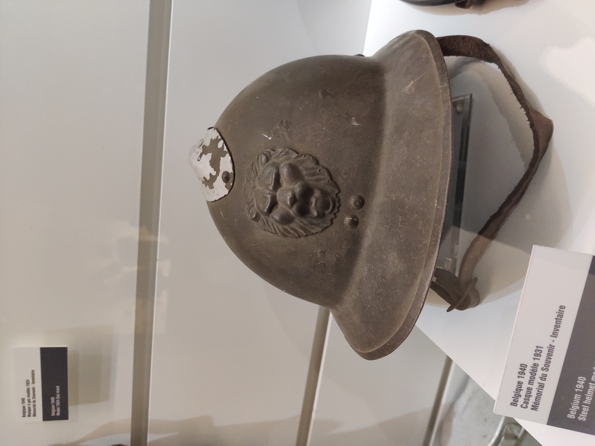 The image displays a close-up view of a historical military helmet, likely from Belgium, dating back to 1940. The helmet is made of dark metal and features a decorative lion's head emblem on the front. It has a wide brim and a leather chin strap that is partially visible. The helmet is positioned on a white surface, which appears to be a display pedestal within a museum or exhibition. In the background, a white wall and a horizontal metallic bar are visible. Two informational labels are also present: one in French, identifying the object as "Belgique 1940 Casque modèle 1931 Mémorial du Souvenir - Inventaire," and another in English, stating "Belgium 1940 Steel helmet mod." The specific location in the city of Dunkirk, France, is not depicted or derivable from the image content.