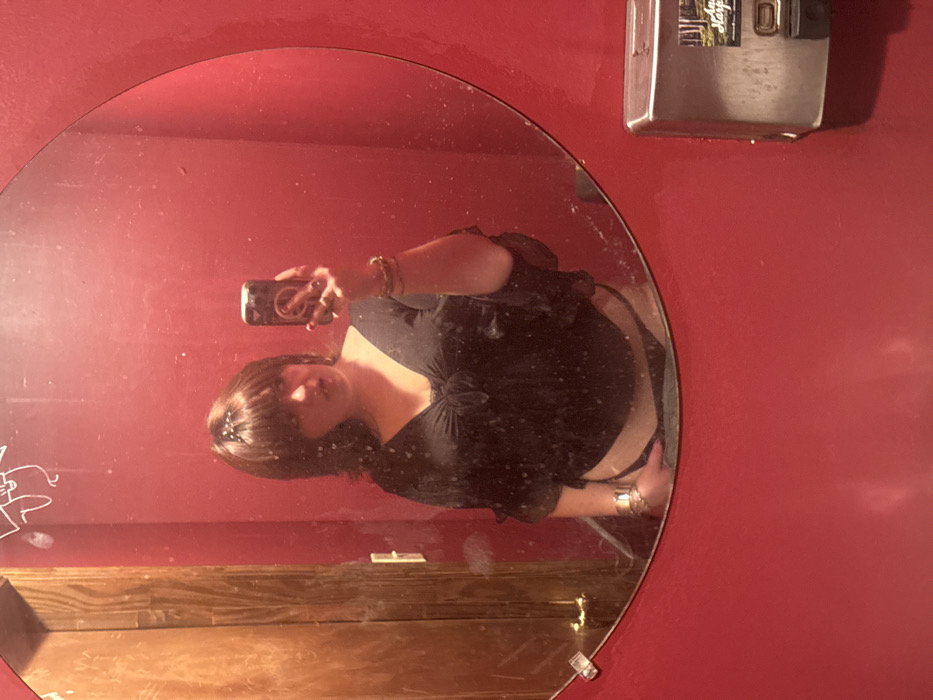 A selfie taken in a round mirror shows a woman with dark hair and bangs, wearing a black shirt and a thin black strap visible on her hip. The mirror is hung on a red wall, with a metal dispenser to the upper right. The lower portion of the mirror reflects wood paneling. Graffiti is visible on the lower-left side of the mirror. The woman holds a phone, reflecting a light source, to take the picture.