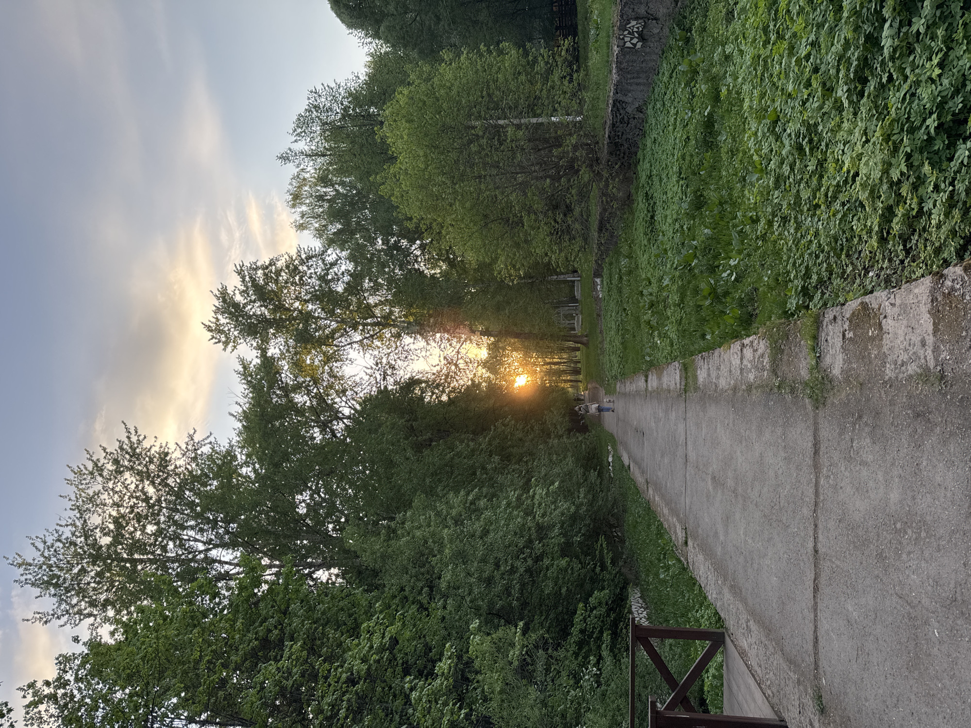 The image shows a concrete path in Izhevsk, Russia, flanked by dense green vegetation. The path is bordered by a low concrete wall on one side and a wooden railing on the other. Sunlight streams through trees at the end of the path. Two figures walk in the distance along the path. The sky is partially cloudy.