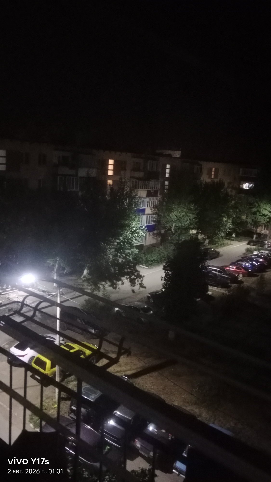 An elevated, wide-angle nighttime photograph captures a residential area in Rudny, Kazakhstan, likely a typical Soviet-era apartment building complex. The scene is dimly lit by a street lamp on the left, illuminating a portion of a paved courtyard with several parked cars. The building, a multi-story concrete structure, is visible in the background with some windows lit from within. Trees are scattered throughout the courtyard, their leaves catching the artificial light. In the foreground, the dark silhouette of a balcony railing or fence partially obscures the view of the street and parked vehicles below. The timestamp in the lower left corner indicates the image was taken on August 2, 2026, at 01:31. No people are visible in the frame.