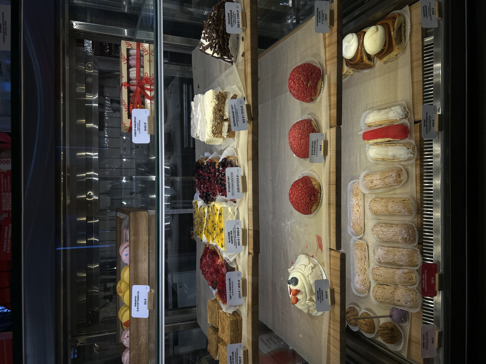 This media file captures a well-stocked dessert display case in what appears to be a bakery or cafe in Saint Petersburg, Russia. The case features multiple shelves filled with various cakes and pastries, meticulously arranged for sale.
On the far left, behind glass, two boxes of macarons are visible. One, adorned with a red ribbon and the text "Набор Макарон И ТЫ ВО ФРАНЦИИ" (Macaron Set AND YOU ARE IN FRANCE), is priced at 649 ₽. Below it, a wooden box holds individual macarons of different colors, labeled "Макарон в ассортименте" (Macaron assorted) for 99 ₽.
The central shelves showcase an array of cake slices and tarts. These include "Медовик шоколадный" (Chocolate Honey Cake) at 199 ₽ per slice (2380 ₽ for a whole cake), "Морковный торт" (Carrot Cake) for 189 ₽ (1510 ₽ for whole), "Ягодный тарт" (Berry Tart) for 209 ₽ (1254 ₽ for whole), "Чизкейк Маракуйя" (Passion Fruit Cheesecake) at 199 ₽ (1990 ₽ for whole), "Тарт с малиной" (Raspberry Tart) for 269 ₽ (1615 ₽ for whole), and "Медовик классический" (Classic Honey Cake) for 199 ₽.
On the right side, a variety of individual pastries are displayed. Red choux buns, labeled "Булочка шу клубничная" (Strawberry Choux Bun), cost 119 ₽. Large buns with white frosting are visible, identified as "Булка маковая в глазури" (Poppy Seed Bun in Glaze) for 139 ₽, and another with cinnamon ("сколицей") for 389 ₽. A selection of white and red-glazed eclairs ("Эклер в ассортименте") are priced at 149 ₽. Below them, additional light brown eclairs dusted with powder are present, along with smaller pastries tagged "Эклер «58» в ассортименте" for 58 ₽. A Pavlova meringue dessert with fresh berries ("Павлова со свежими ягодами") is priced at 349 ₽. Cake pops ("Кейк-попс") are also displayed for 99 ₽.
Visible text on the display also includes encouraging phrases like "ОЧЕНЬ ВКУСНО!" (VERY TASTY!) and "ОСТОРОЖНО!" (CAUTION!). The pricing is consistently in Russian Rubles (₽). No people are visible in the image.