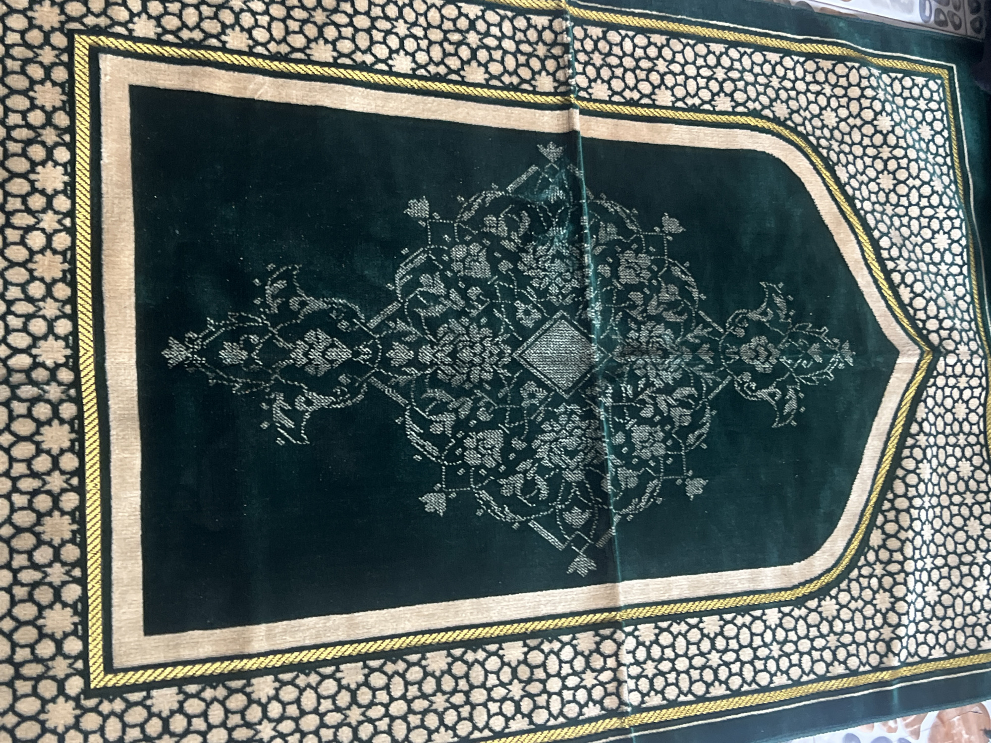 The image shows a dark green prayer rug with an ornate, symmetrical silver-colored design at its center. The rug has a tan border outlined with gold, and a patterned outer edge of repeating black and white shapes. Given the nature of a prayer rug, the image potentially originates from a Muslim household, and the city mentioned, Minna, is located in Nigeria. No people, actions, or interactions are visible in this image.