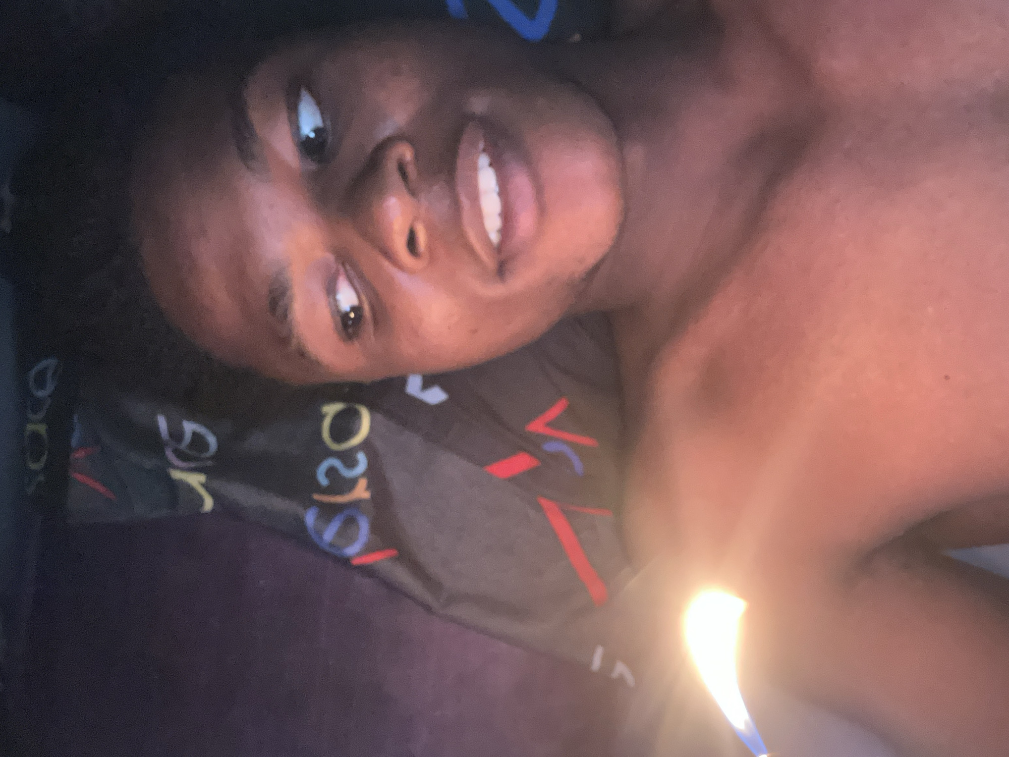 The image is a close-up, low-angle selfie of a young Black male lying down, possibly on a bed, in a dimly lit room. The subject is looking to his right, with a slight smile. He is shirtless, and a bright light source, possibly a phone screen or a small flame, is positioned on the left side of the frame, casting a strong glare. The background shows a dark fabric with colorful, stylized text and geometric shapes. Based on the provided context, this scene is set in the city of Ihiagwa, Nigeria.