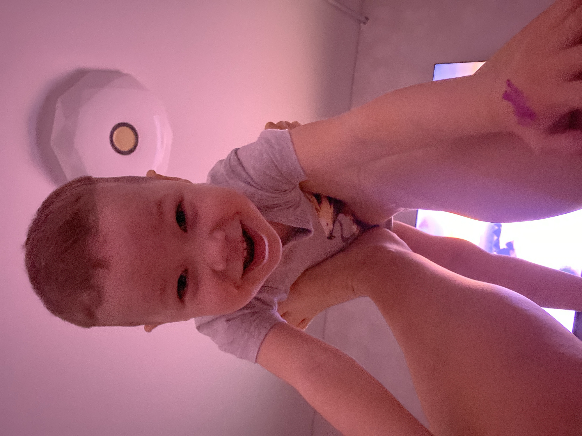 A low-angle close-up shot shows a young child being held up by an adult. The child, likely a toddler, is wearing a gray t-shirt and is smiling broadly, revealing their teeth. Above the child's head, a round ceiling light fixture is visible. The adult's arms, pale in complexion, are wrapped around the child. On the adult's arm there is a purplish mark. Behind the adult's arm, part of a lit television screen is visible. The scene appears to be indoors, possibly a home interior. It is not possible to pinpoint the location as the city of Sochi, Russia, without further visible details or context.