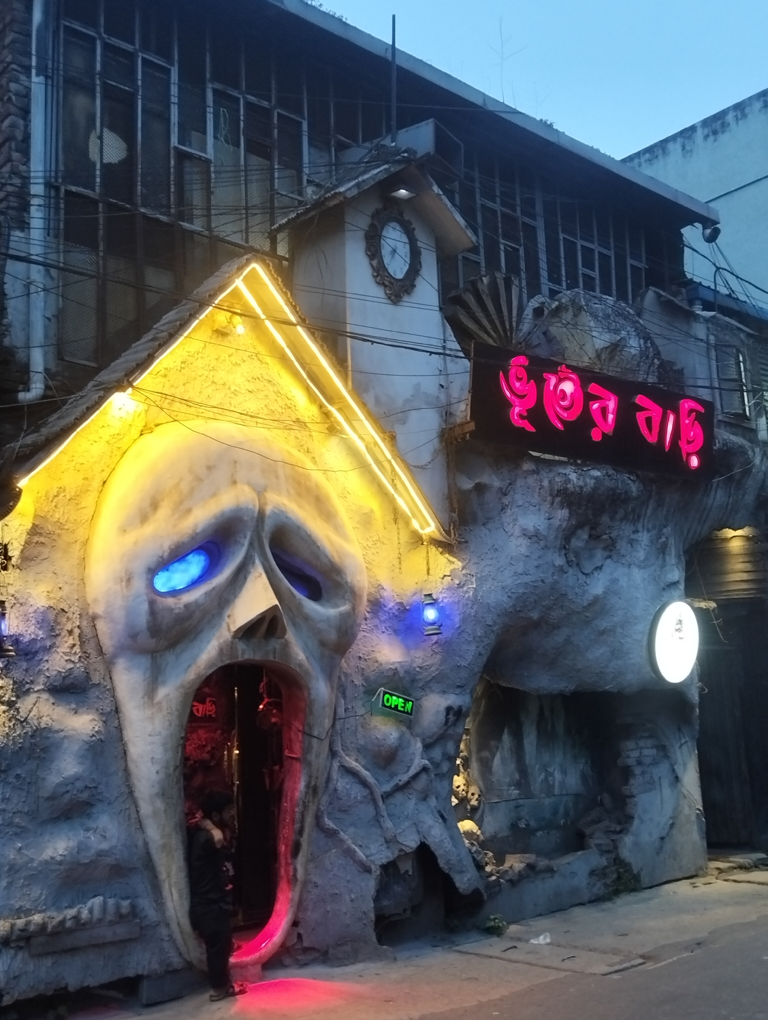 The image displays the eerie and dramatic entrance of a horror-themed establishment, likely a "Haunted House," observed during dusk or evening hours. The prominent feature is a massive, sculpted facade resembling a pale, gaunt face or skull with a tormented expression. Its eyes glow with a piercing blue light, while its wide-open mouth, illuminated in a deep red, serves as the primary entrance.
Above this monstrous face, yellow neon-like lights delineate a triangular roof structure, drawing attention to the entrance. To the right of the main entryway, a dark archway or cave-like passage is visible, featuring several skull-like objects and a circular white light mounted on the wall. A black sign with vibrant pink Bengali text, reading "ভূতের বাড়ি" (Bhuter Bari), which translates to "Ghost House" or "Haunted House," is prominently displayed above this section, explicitly stating the venue's theme. A small green "OPEN" sign is also visible near the entrance, indicating that the establishment is welcoming visitors.
One person, clad in dark clothing, is seen standing just inside the red-lit mouth entrance, partially obscured. Above the themed facade, an older building with multiple windows covered by metal grates is visible, suggesting an urban setting. A small gabled structure with a clock face is also mounted above the central entrance. The overall scene, with its dramatic lighting, macabre decorations, and the lone individual entering, creates an atmosphere of spooky adventure and intrigue, hinting at the thrilling experiences within. The Bengali text strongly indicates a location in a Bengali-speaking region.