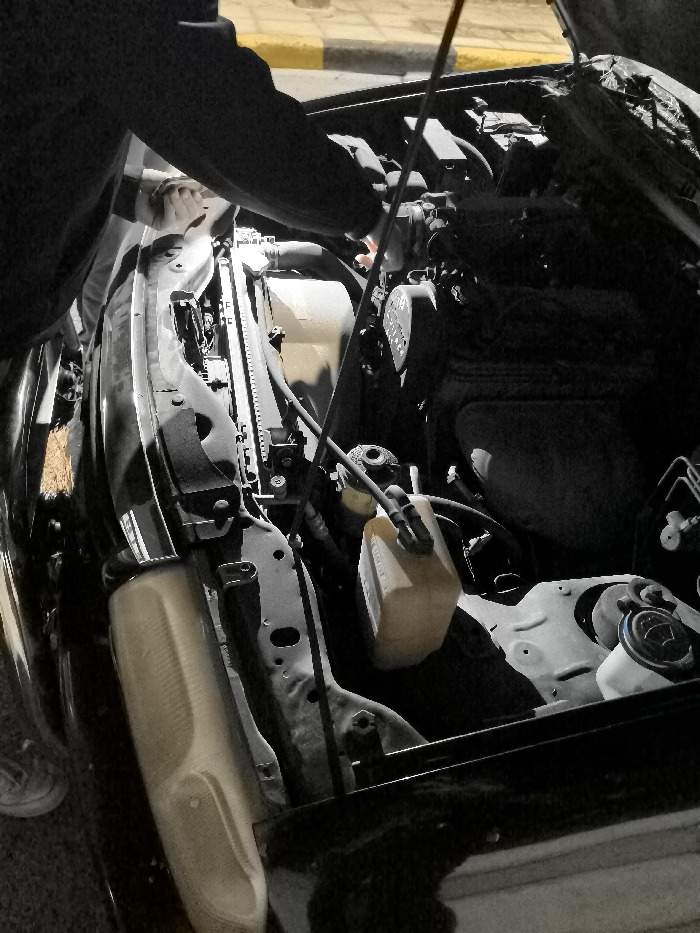The image presents a close-up view of a person engaged in automotive work, likely at night or in low light conditions, given the strong contrasts and shadows. The scene focuses on the open engine bay of a car. A person, visible from the torso and arm in the upper left, leans over the engine, with their right hand seemingly manipulating a dipstick, suggesting they are checking fluid levels such as oil or transmission fluid.
The engine bay reveals various components including a radiator along the left side, numerous hoses, wires, and engine parts. A translucent, light-colored plastic fluid reservoir, possibly for coolant or power steering fluid, is visible on the right side of the engine bay. A portion of a car headlight can be seen at the bottom left of the frame. Faint text reading "ENGINE" is discernible on a component in the upper right section of the engine.
In the background, a yellow-painted curb and a paved road are visible, indicating an outdoor setting, potentially a street or parking area in Iraklio, Greece. The activity portrays a moment of vehicle inspection or maintenance.