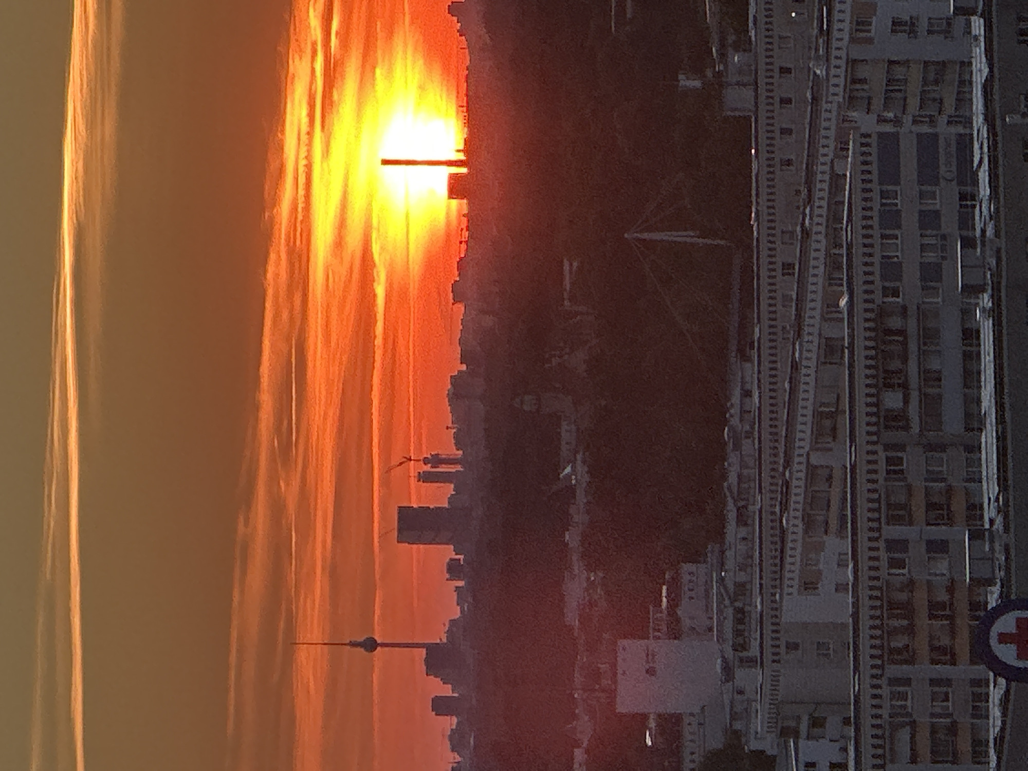 The image captures a city skyline during sunset, with the sun a brilliant orange orb dominating the horizon. The sky is a vibrant gradient of orange, yellow, and streaks of red, with thin clouds adding texture. Silhouetted against the bright sky are various buildings, including a prominent tall tower with a spherical top, resembling the Fernsehturm (TV Tower) in Berlin. Adjacent to it, other skyscrapers and cranes suggest an active construction site within the cityscape. In the foreground, several blocks of residential buildings with uniform balconies and windows are visible. A large, dark area in the mid-ground, likely covered in trees, separates the distant skyline from the closer buildings. The overall scene depicts an urban environment bathed in the warm, dramatic light of a setting sun. The presence of the Fernsehturm strongly suggests the location is Berlin, Germany. There are no people or animals visible in the image.