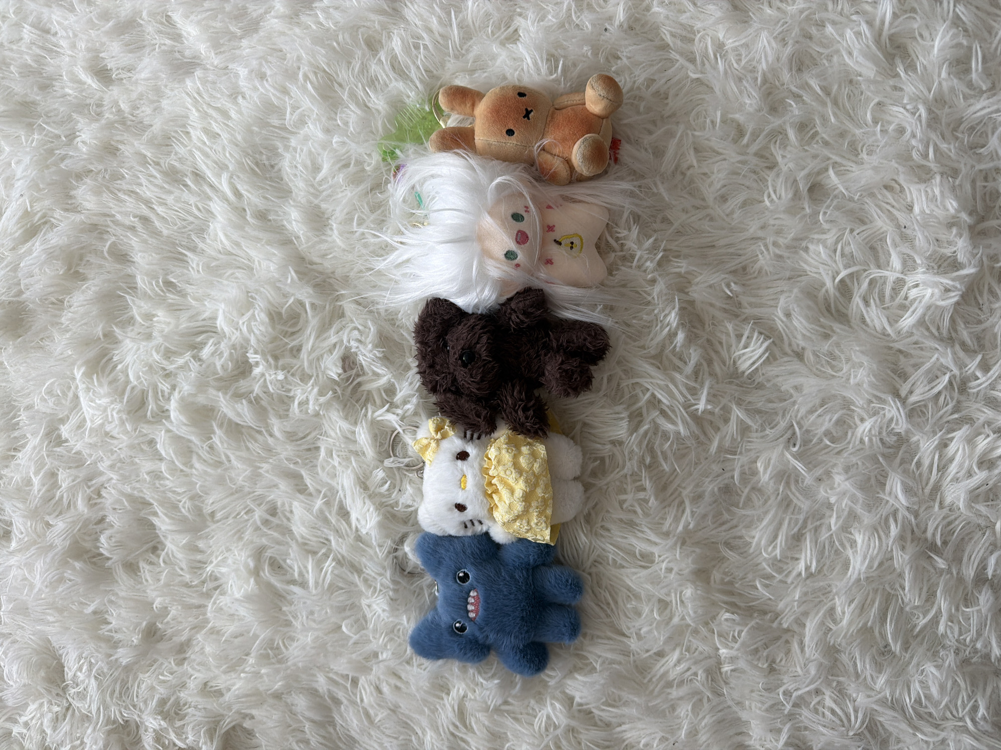 A top-down medium shot shows five small plush toys stacked on a white, shaggy rug. From top to bottom, the toys are: a brown Miffy-like rabbit, a pale-yellow star with white hair, a dark-brown bear, a white Hello-Kitty-like cat with a yellow dress, and a blue shark. The location and city are not discernible from the image.